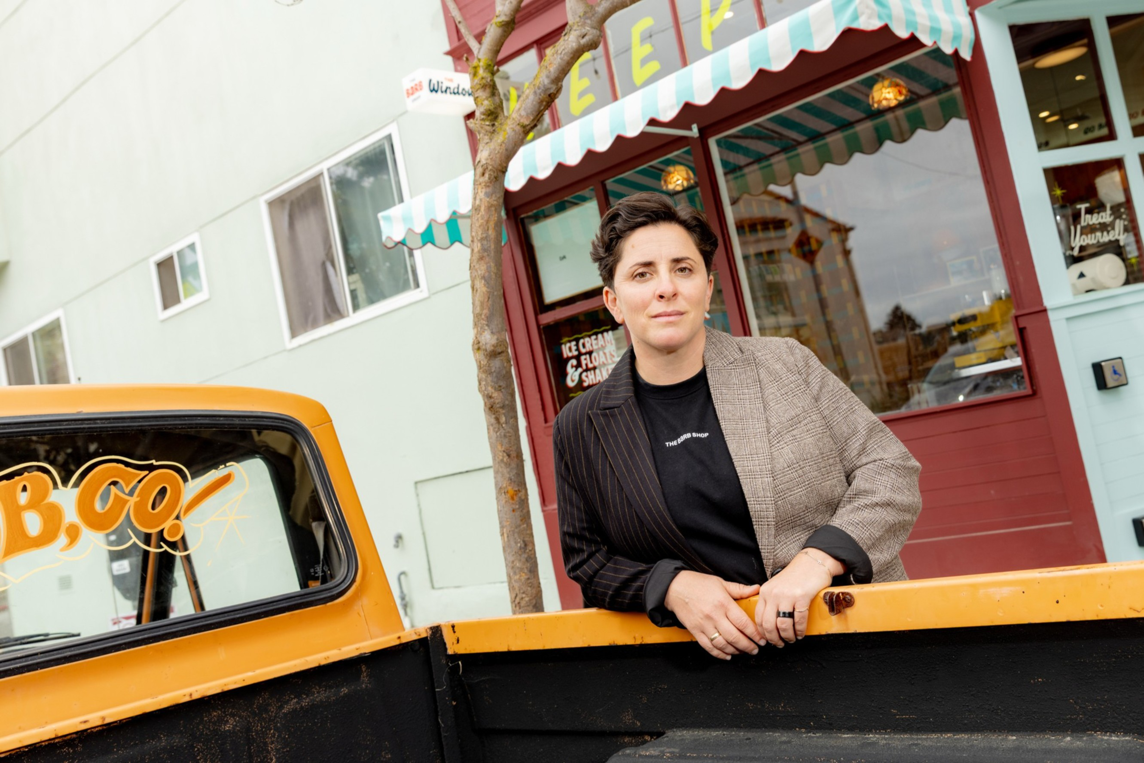 A person in a half-striped, half-plaid jacket leans on an orange truck bed outside a shop with a green and white striped awning.