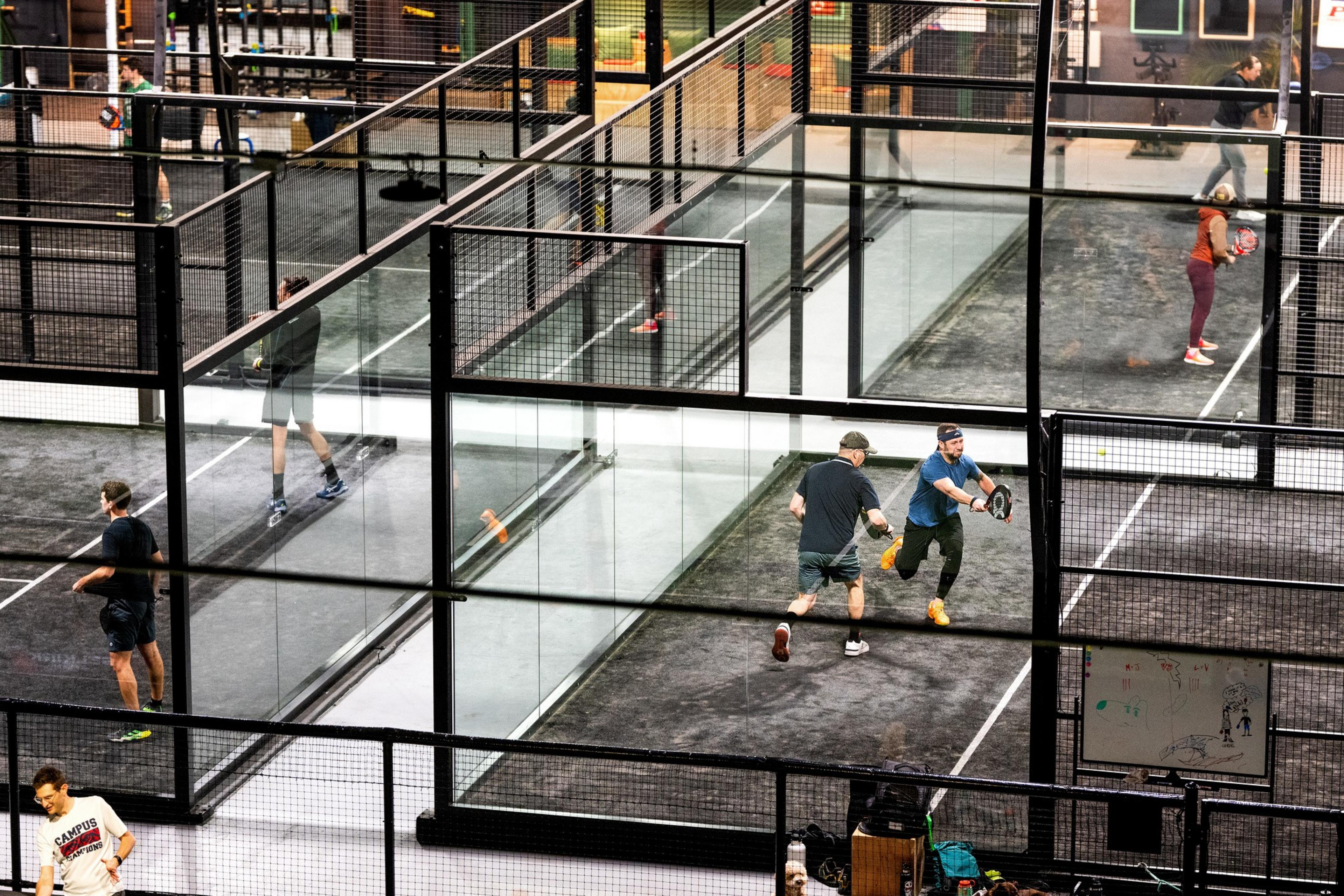 Several people are playing paddle sports on side-by-side indoor courts with glass and net enclosures, all actively engaged in their games.