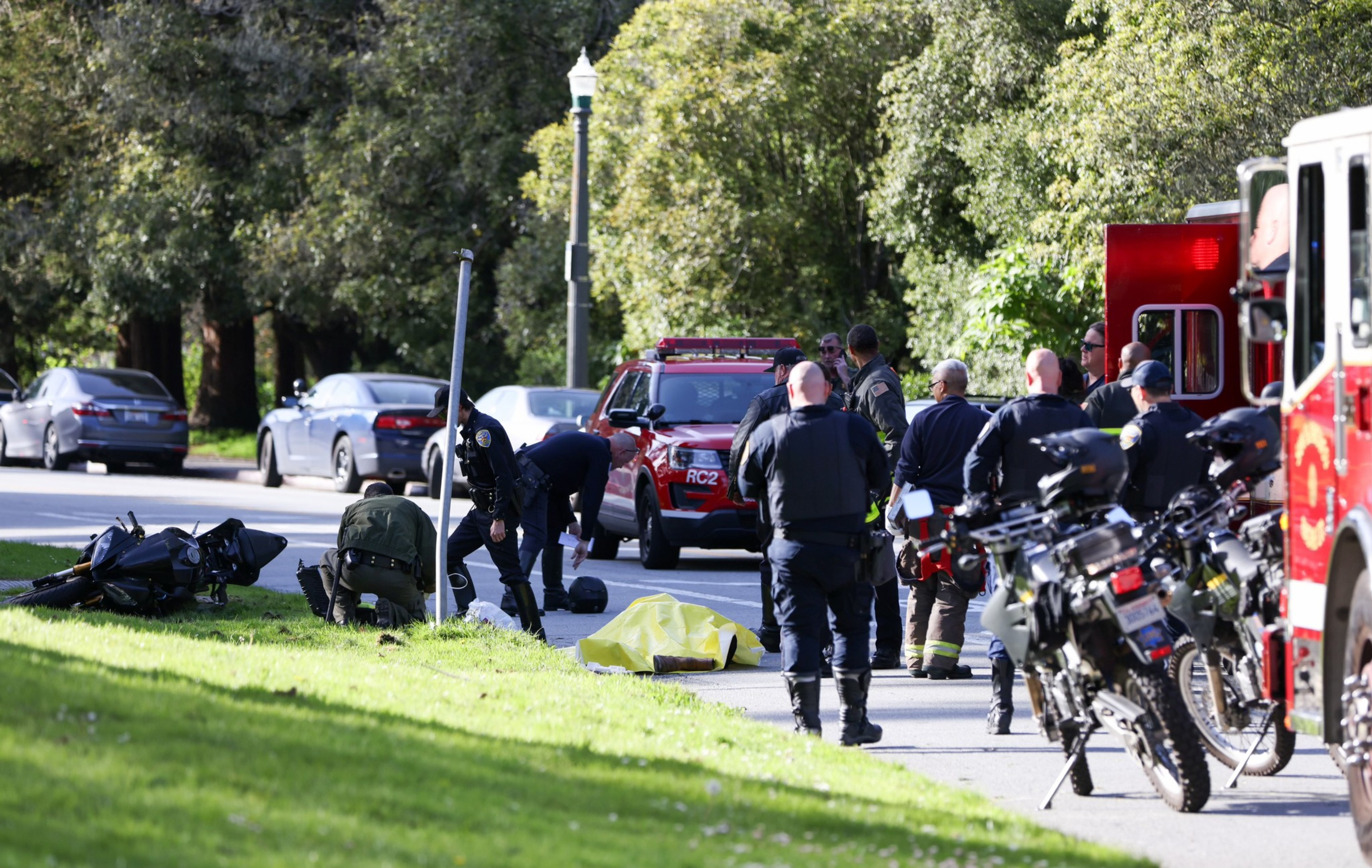 Emergency responders at a park with gear scattered, a yellow tarp on the ground, and vehicles in the background.