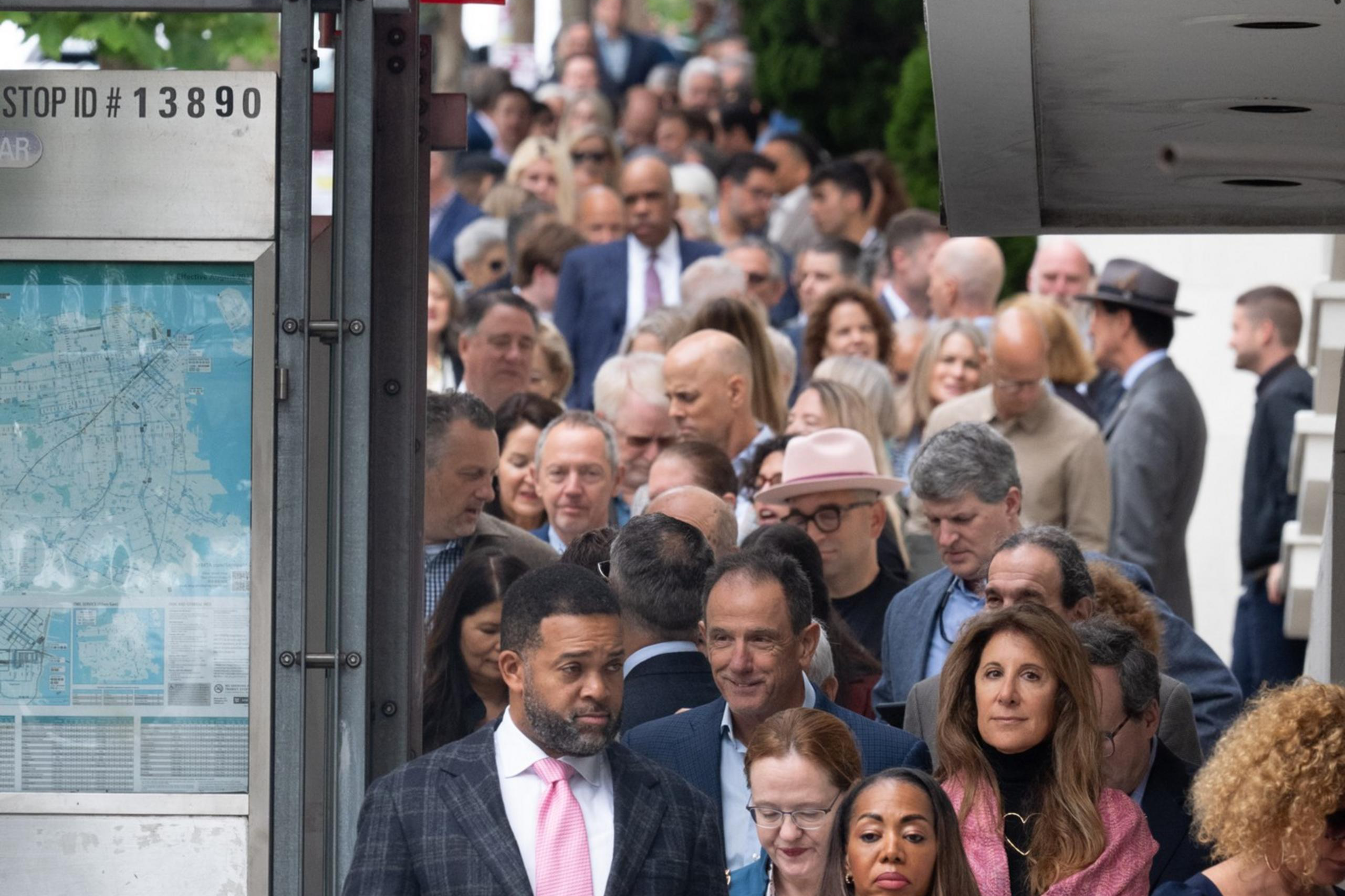 A large, diverse crowd stands in line on a sidewalk, with police officers in uniform visible in the foreground. Many people appear to be dressed formally.