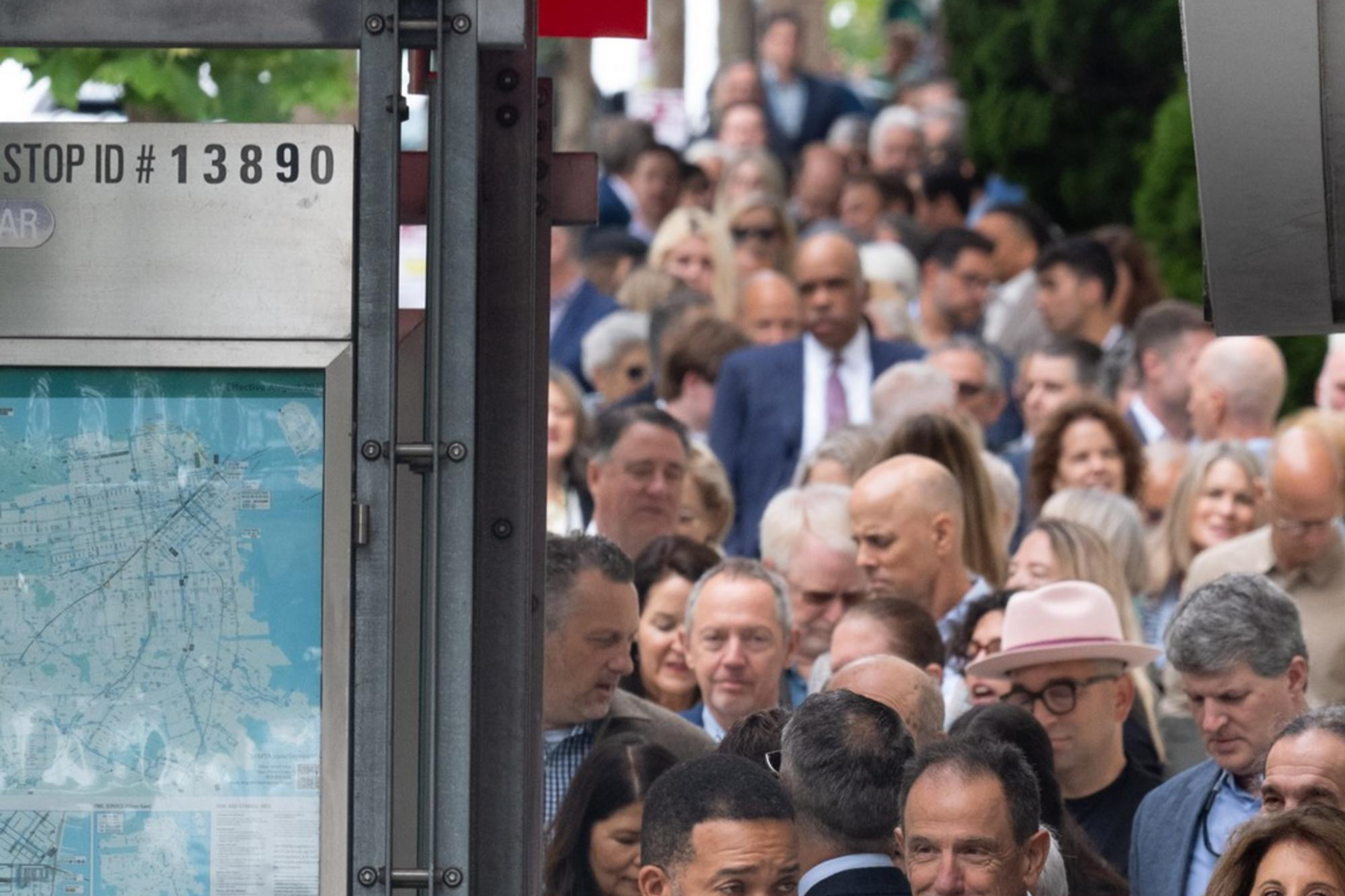 A large, diverse crowd stands in line on a sidewalk, with police officers in uniform visible in the foreground. Many people appear to be dressed formally.