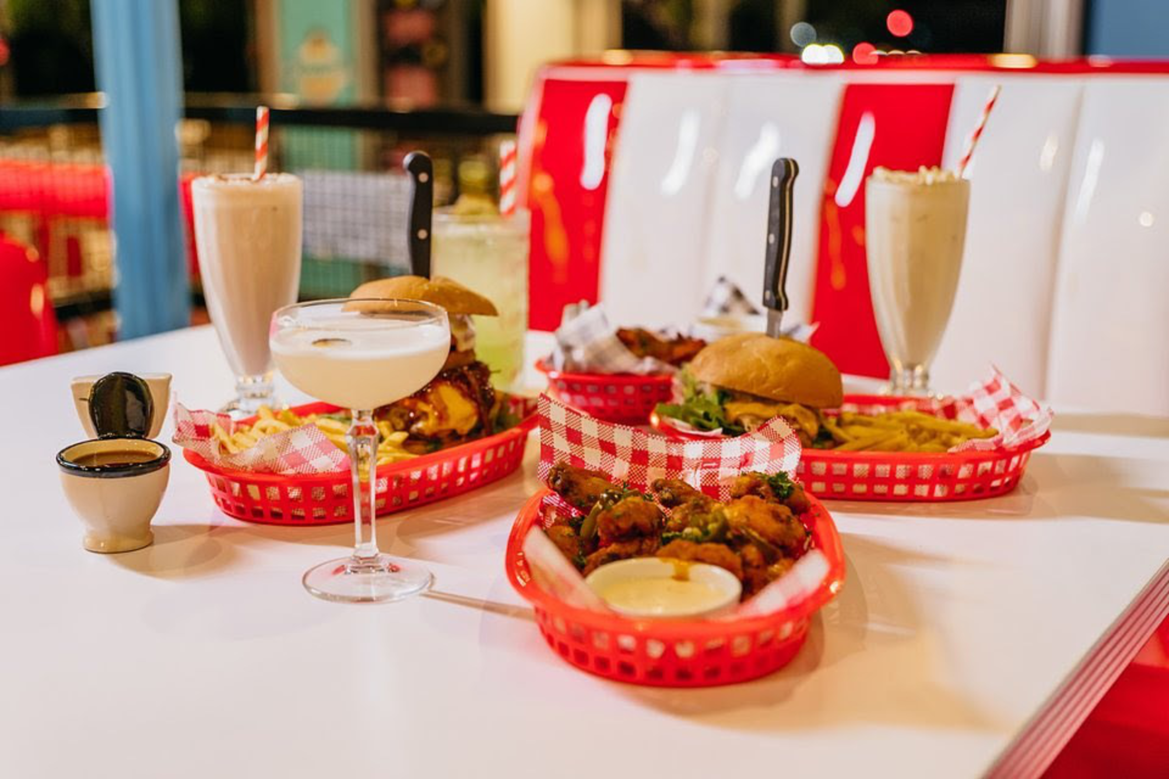 Two cheeseburgers with fries, a basket of wings with dipping sauce, two milkshakes with striped straws, and two cocktails on a white tabletop.