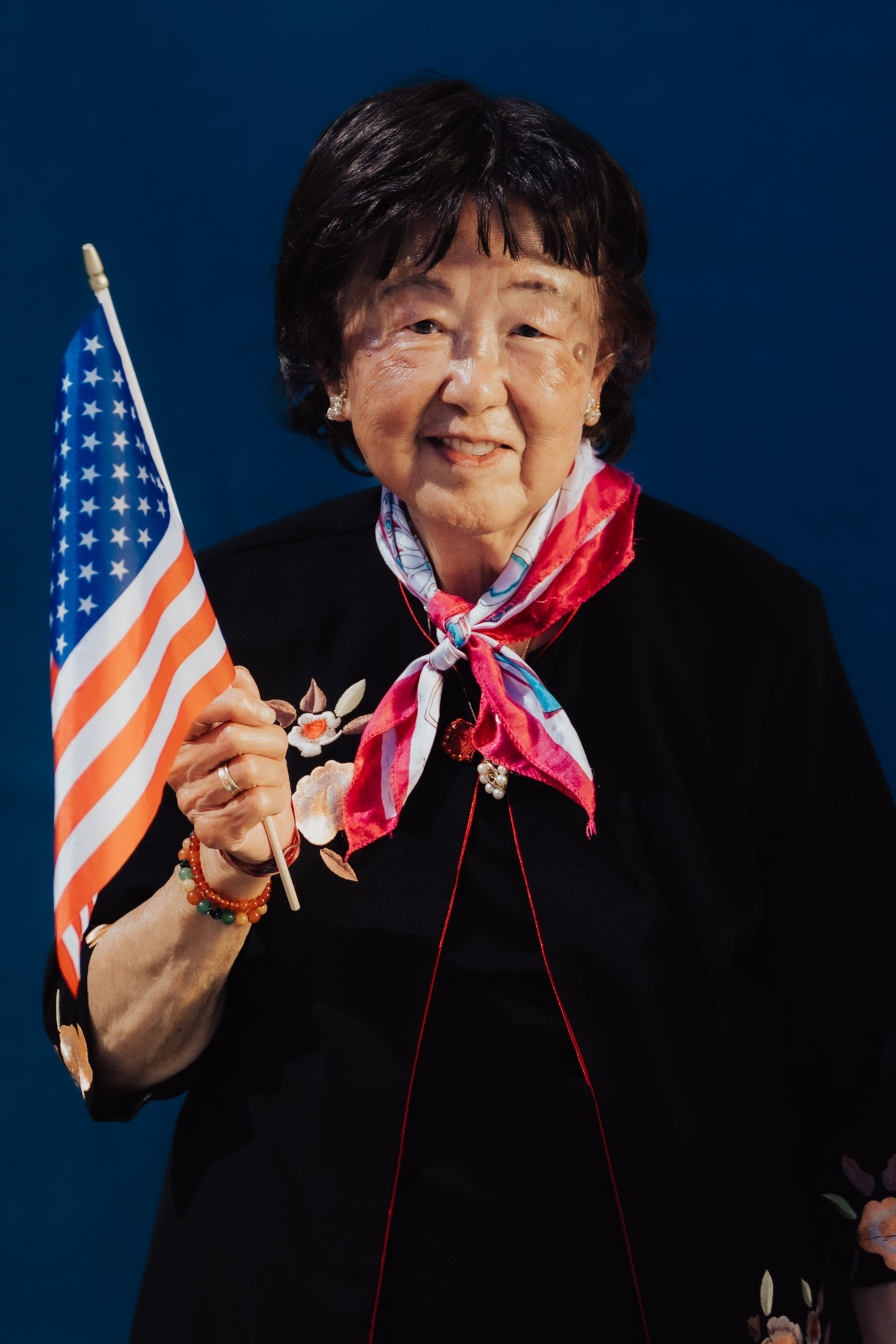 An elderly woman with short dark hair smiles while holding a small American flag, wearing a floral black top and a red, white, and blue scarf.