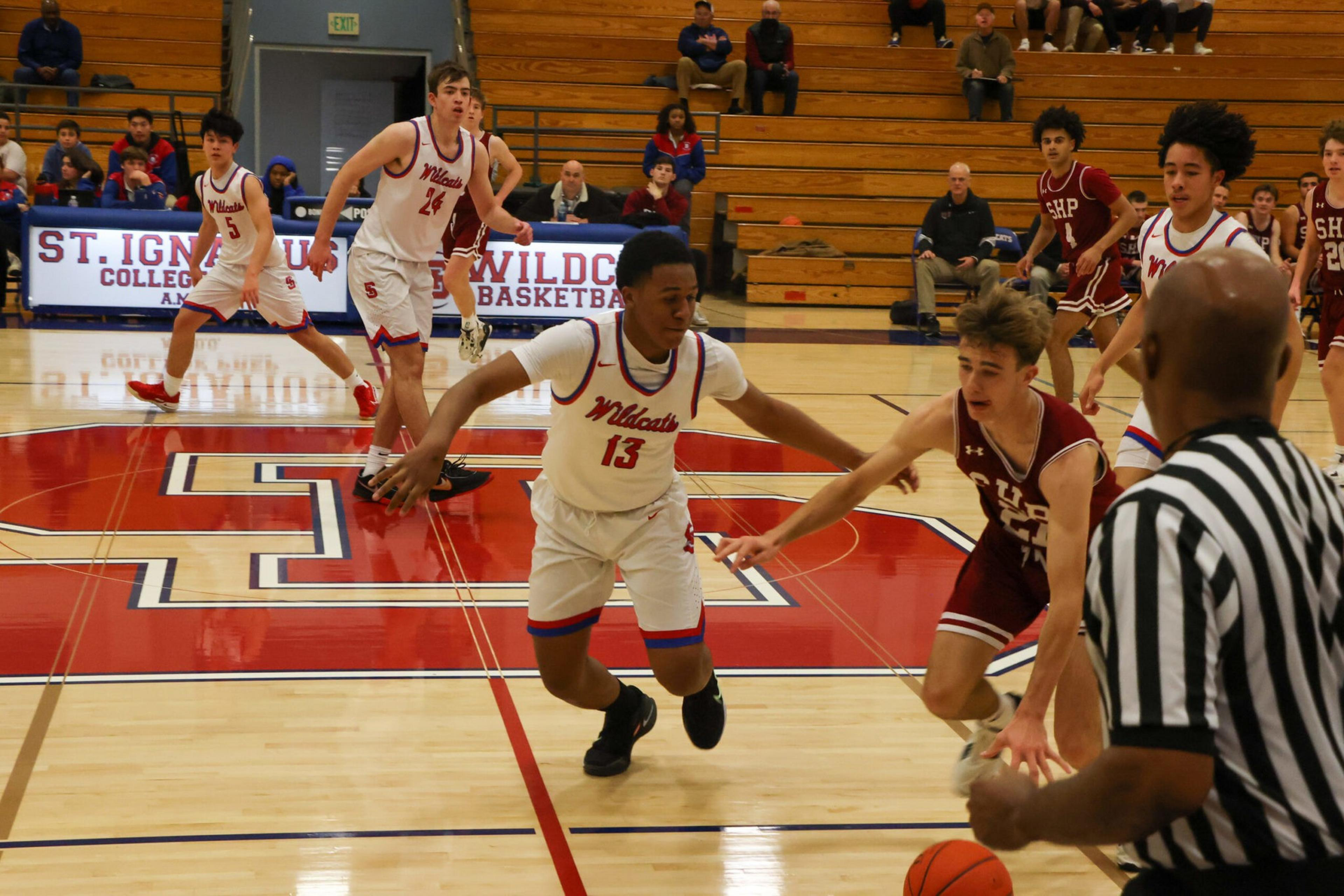 Two basketball players, one in a white Wildcats jersey and one in a maroon SHP jersey, compete near the referee holding the ball on a gym court.