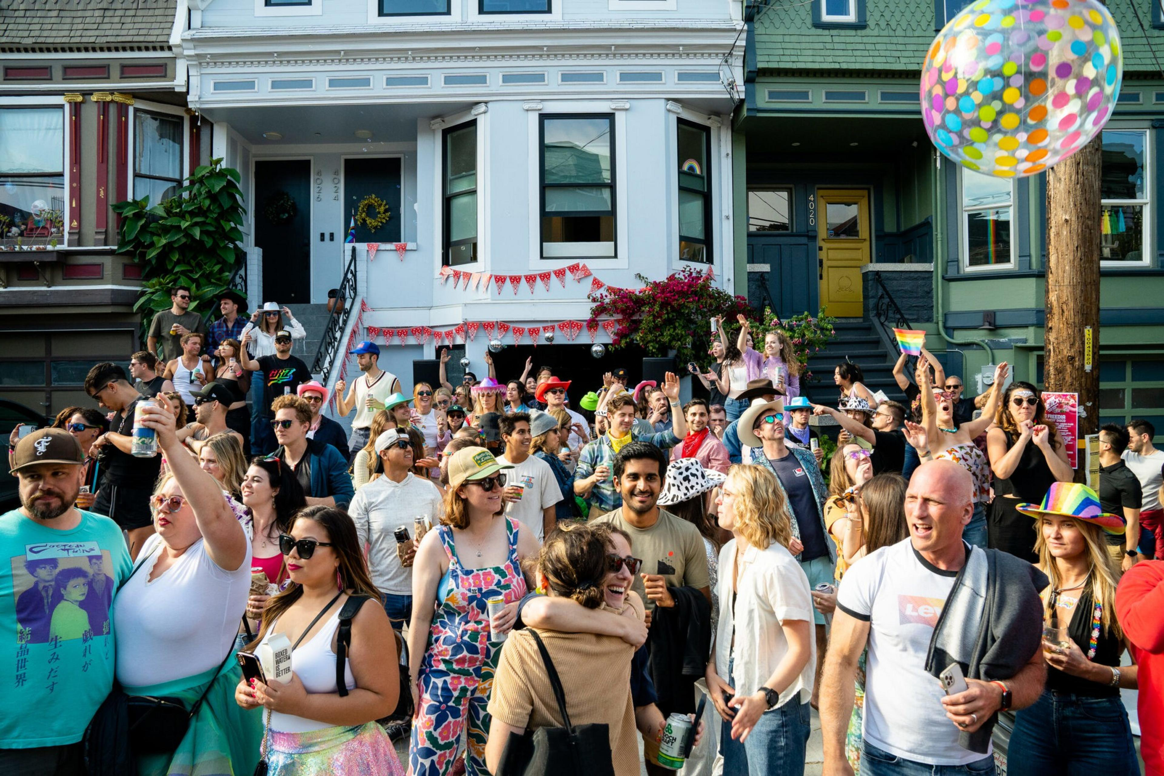A large crowd of diverse people enjoy an outdoor street party, with many holding drinks, some wearing colorful hats, and a large multicolored balloon floating nearby.