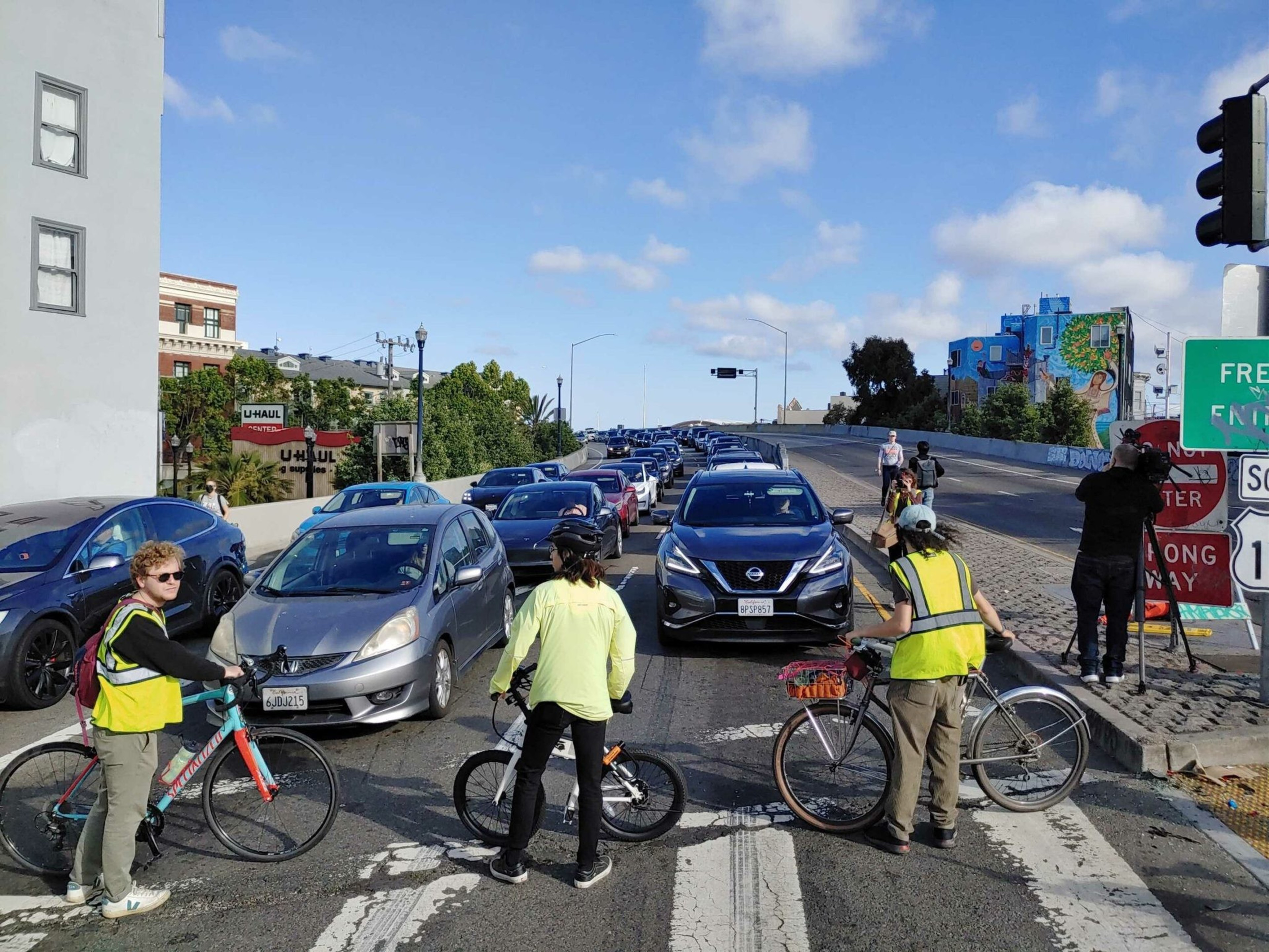 Three cyclists wearing safety vests stand in front of a line of cars stopped at a traffic light on a sunny day.