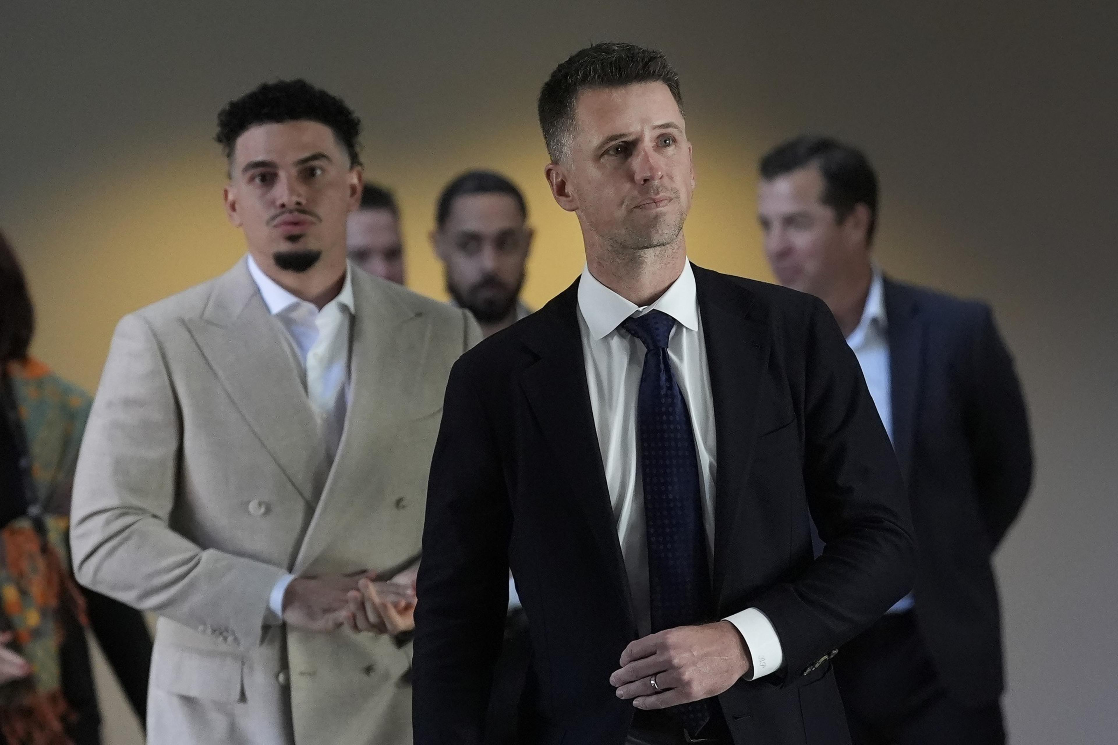 A group of men in suits stand together. The foreground shows two men, one in a light suit, the other in a dark suit and tie, both with serious expressions.