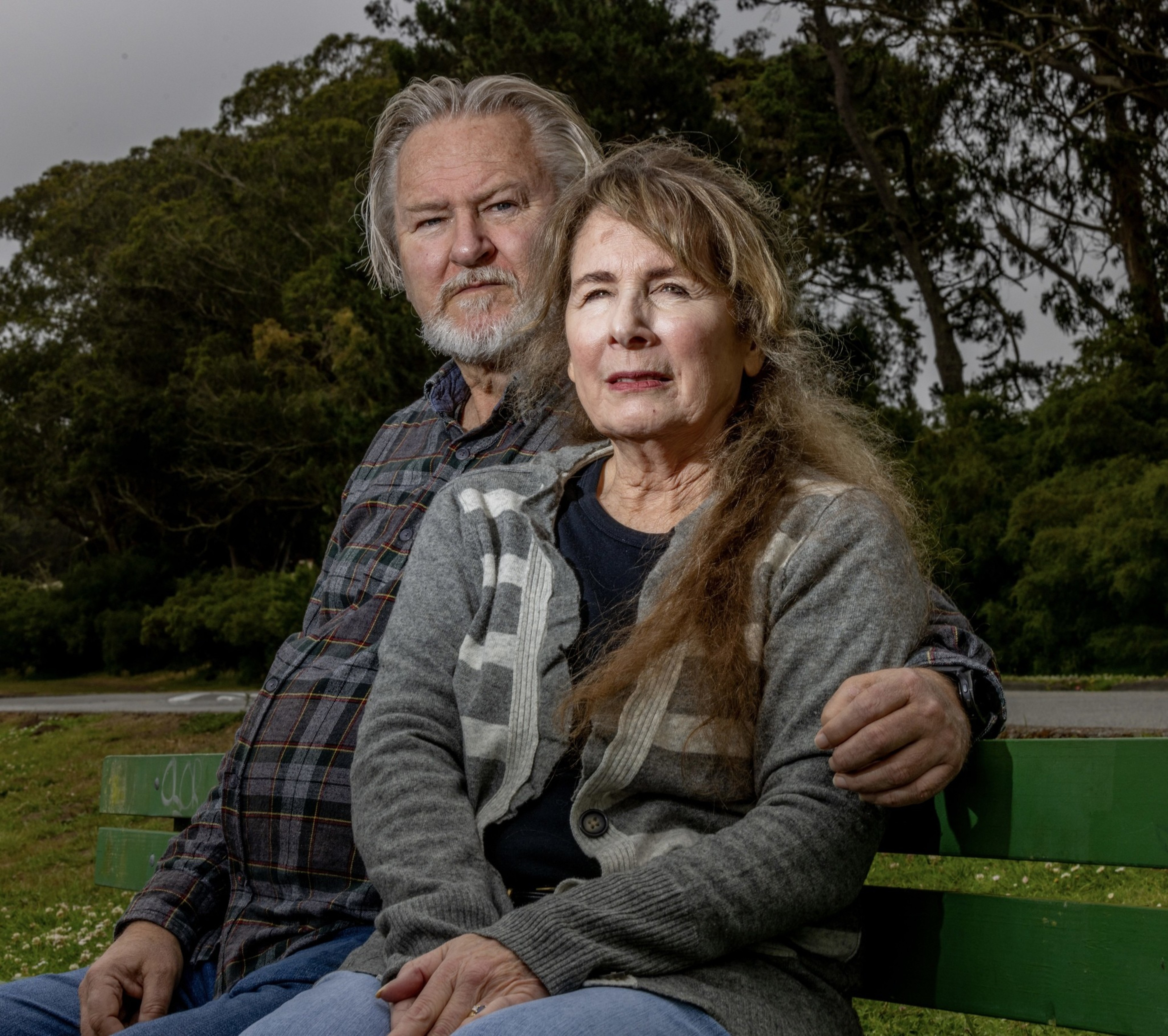 A mature couple sits closely on a green bench outdoors, surrounded by lush trees; the man has gray hair and a beard and wears a plaid shirt, while the woman has long hair and a gray cardigan.