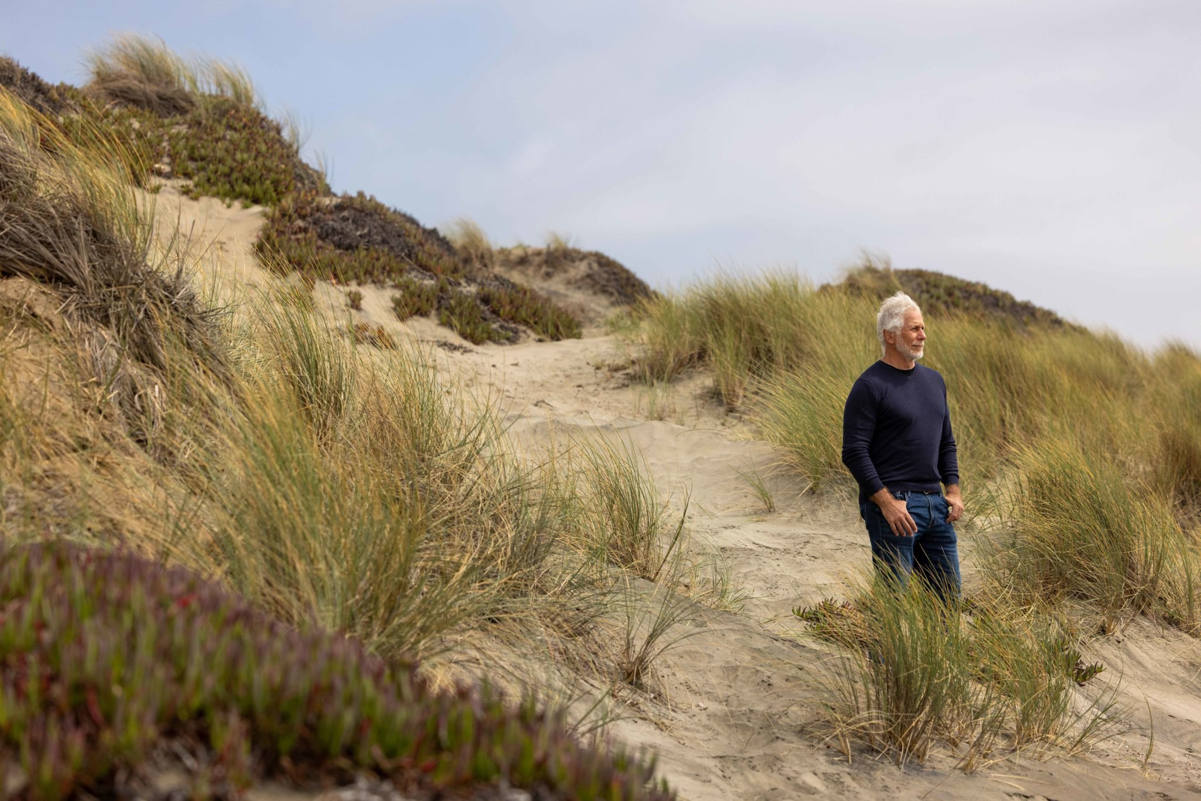 An older man with gray hair and a beard stands on sandy dunes covered with tall grass, looking thoughtfully into the distance.