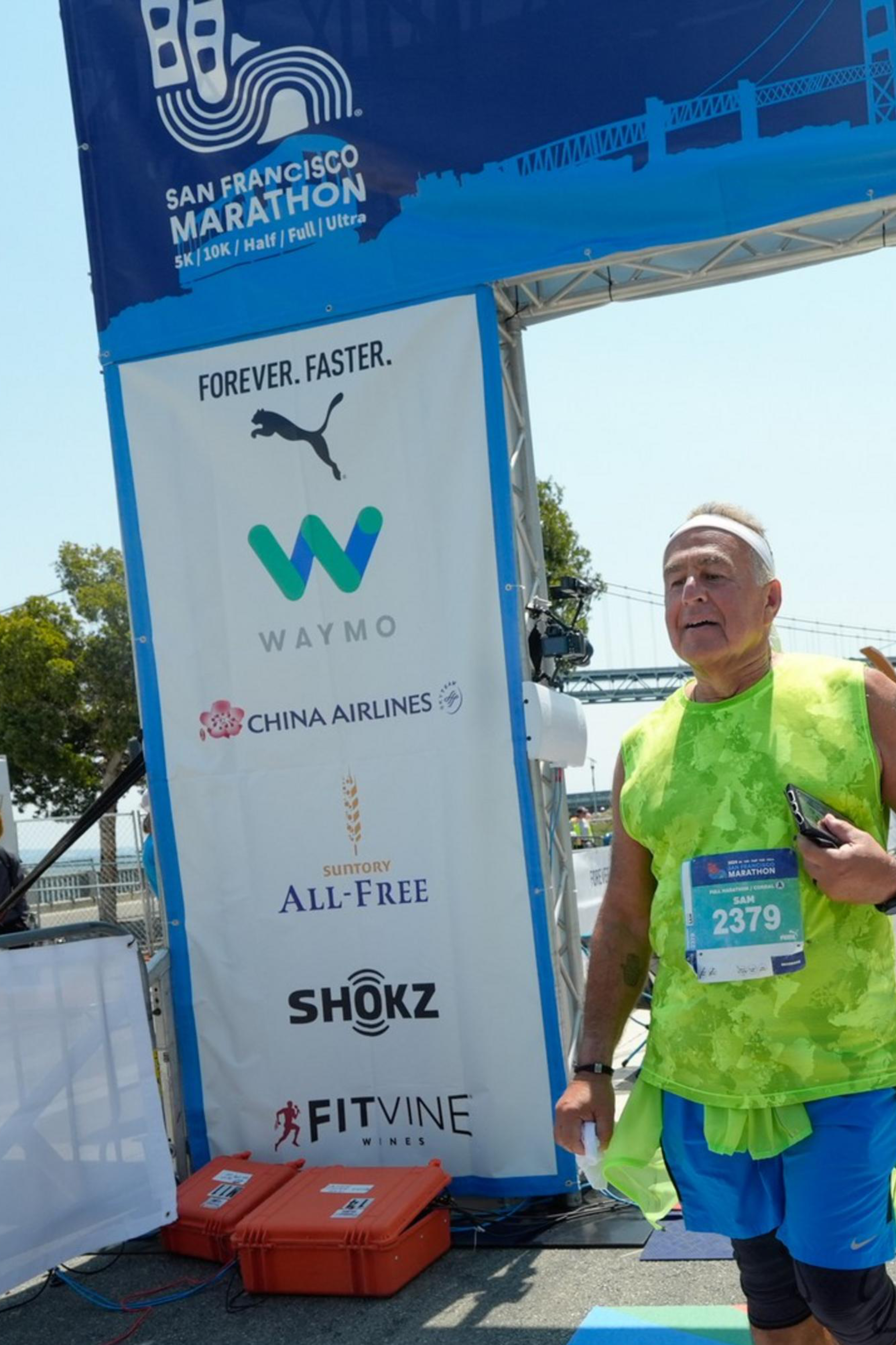A runner wearing a green shirt crosses the finish line at the San Francisco Marathon under a blue arch with sponsor logos and a timing clock overhead.
