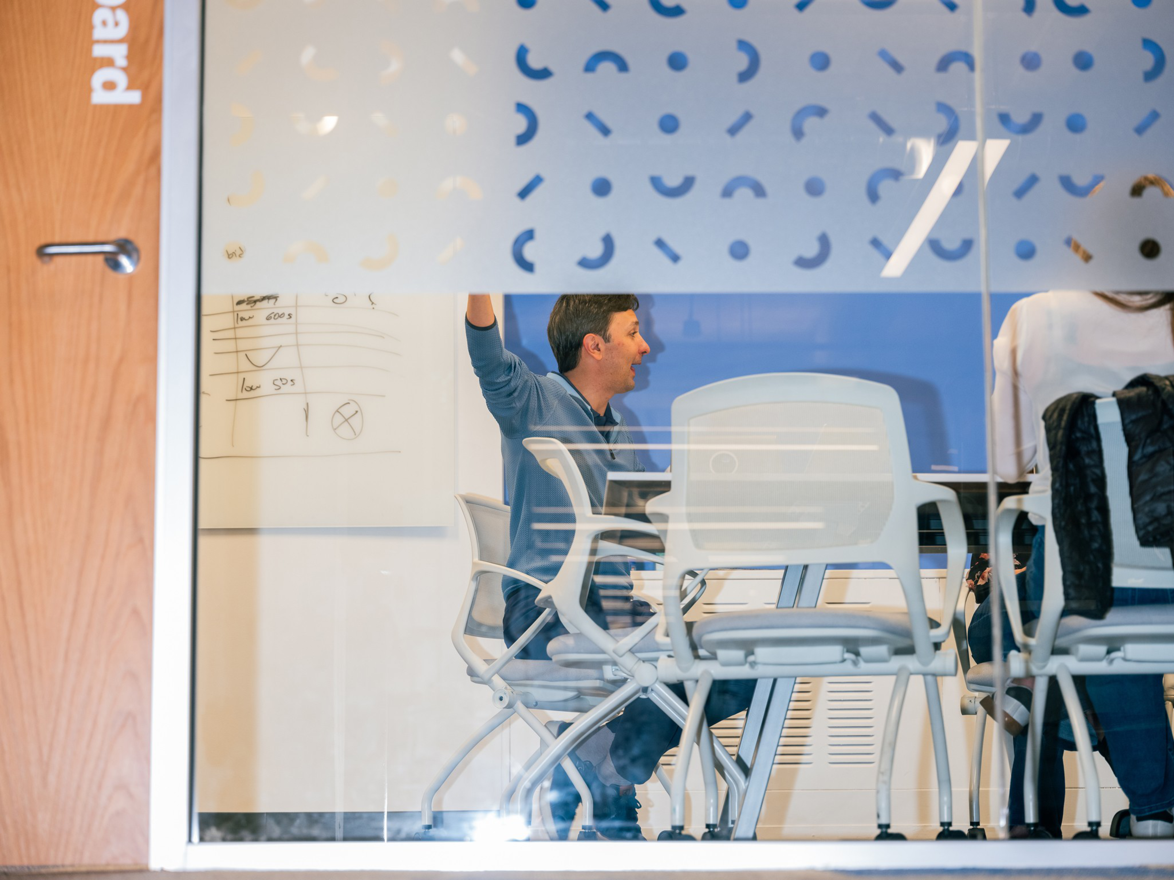 A man in a blue shirt raises his hand while sitting in a meeting room with white chairs and a whiteboard behind him. Another person sits nearby.