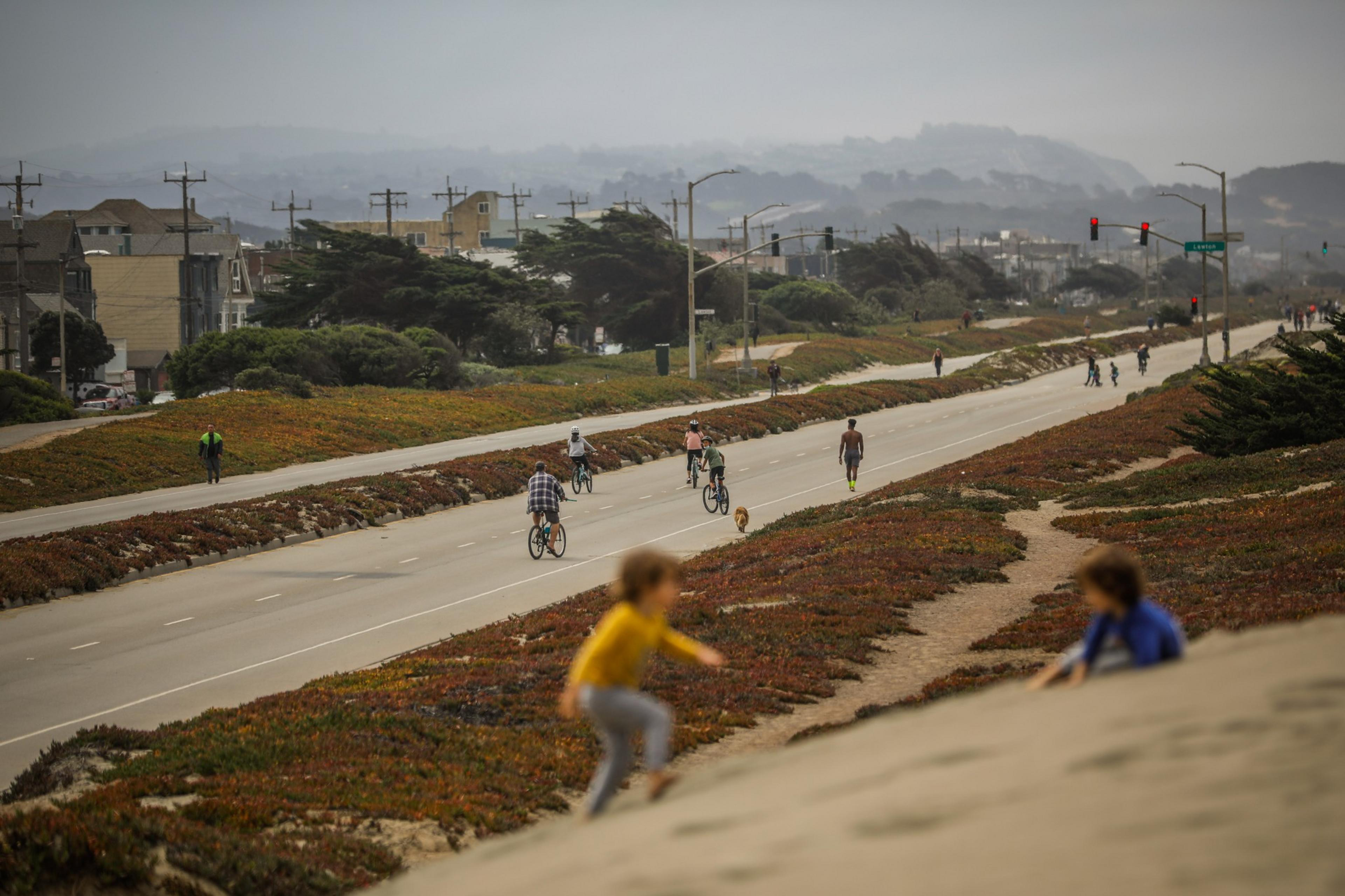 People walk, bike, and jog along a wide, empty road with greenery on both sides, while two children play on a sandy embankment in the foreground.