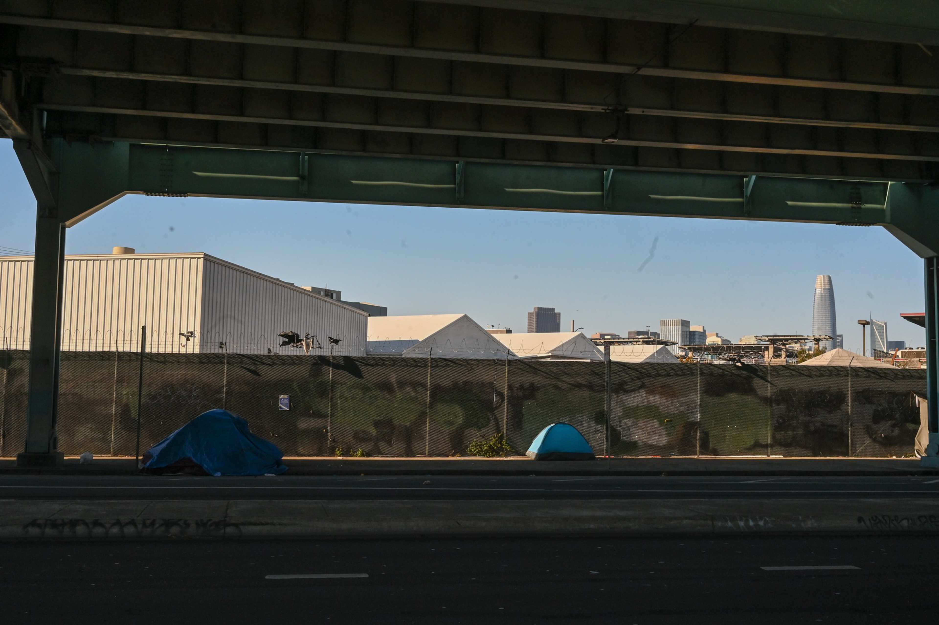 Two small tents are set up under a green highway overpass, with a chain-link fence and industrial buildings in the background and a city skyline in the distance.