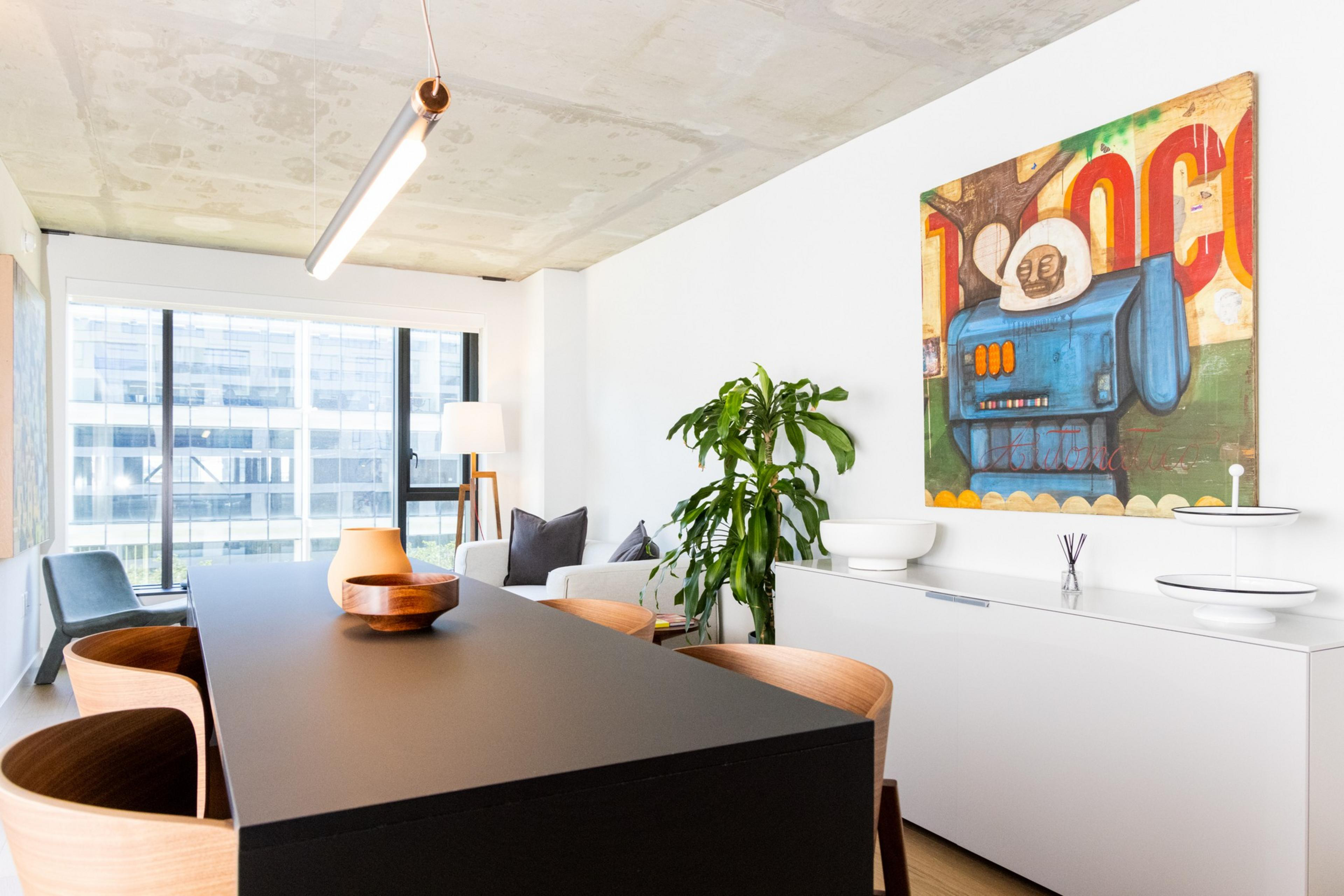 A modern living room with wooden chairs around a black table, a large window, a plant, a white cabinet, and a vibrant painting on a white wall.