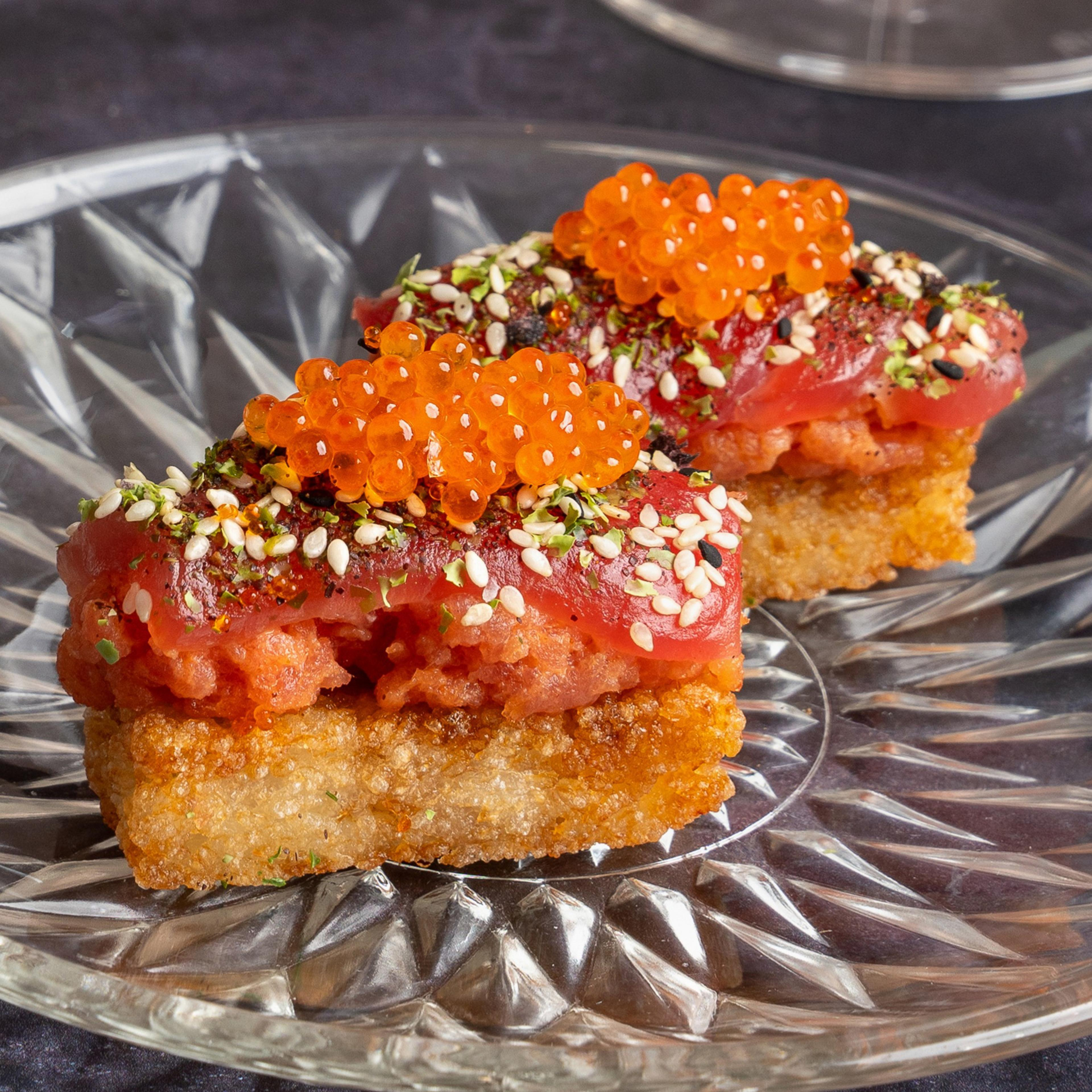 Two pieces of sushi with crispy rice base, topped with spicy tuna, orange roe, and garnished with sesame seeds and herbs, sit on a clear glass plate.