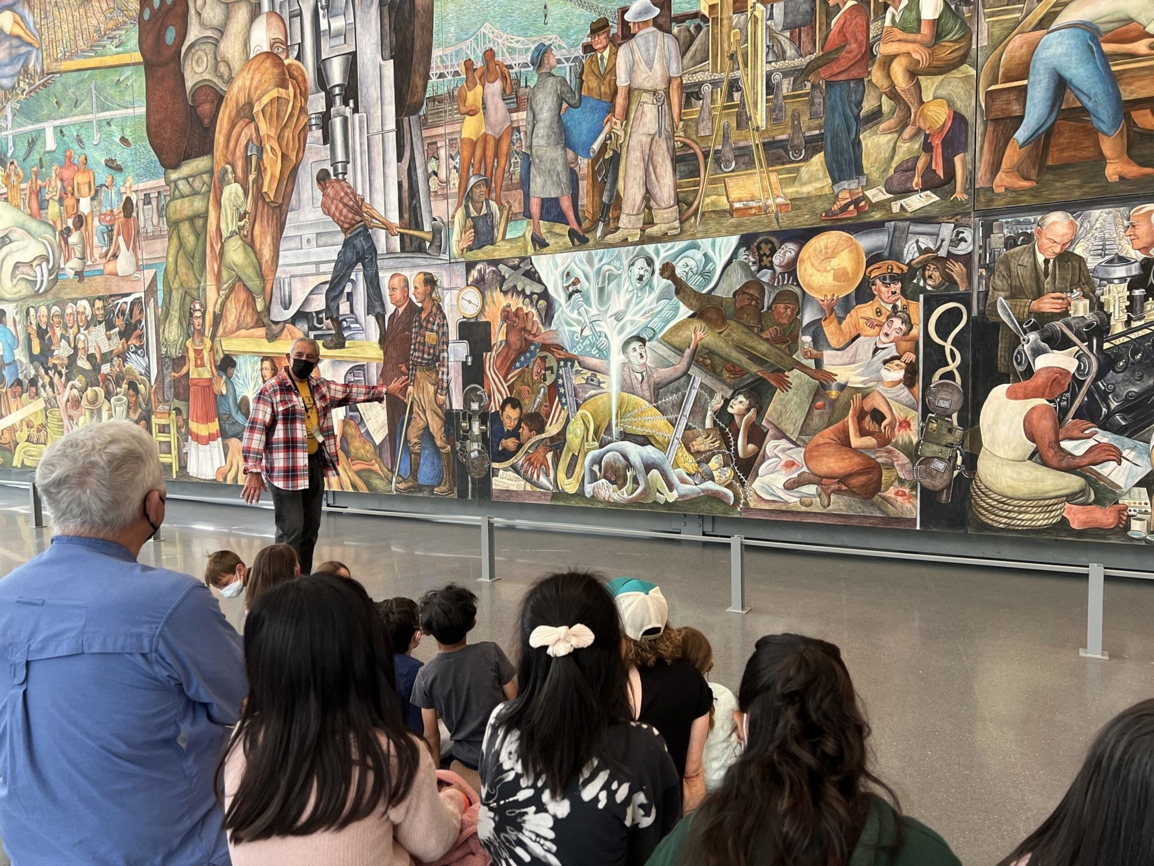 A group of people, including children, sit on the floor listening to a masked guide explaining a large, colorful mural depicting various historical and industrial scenes.