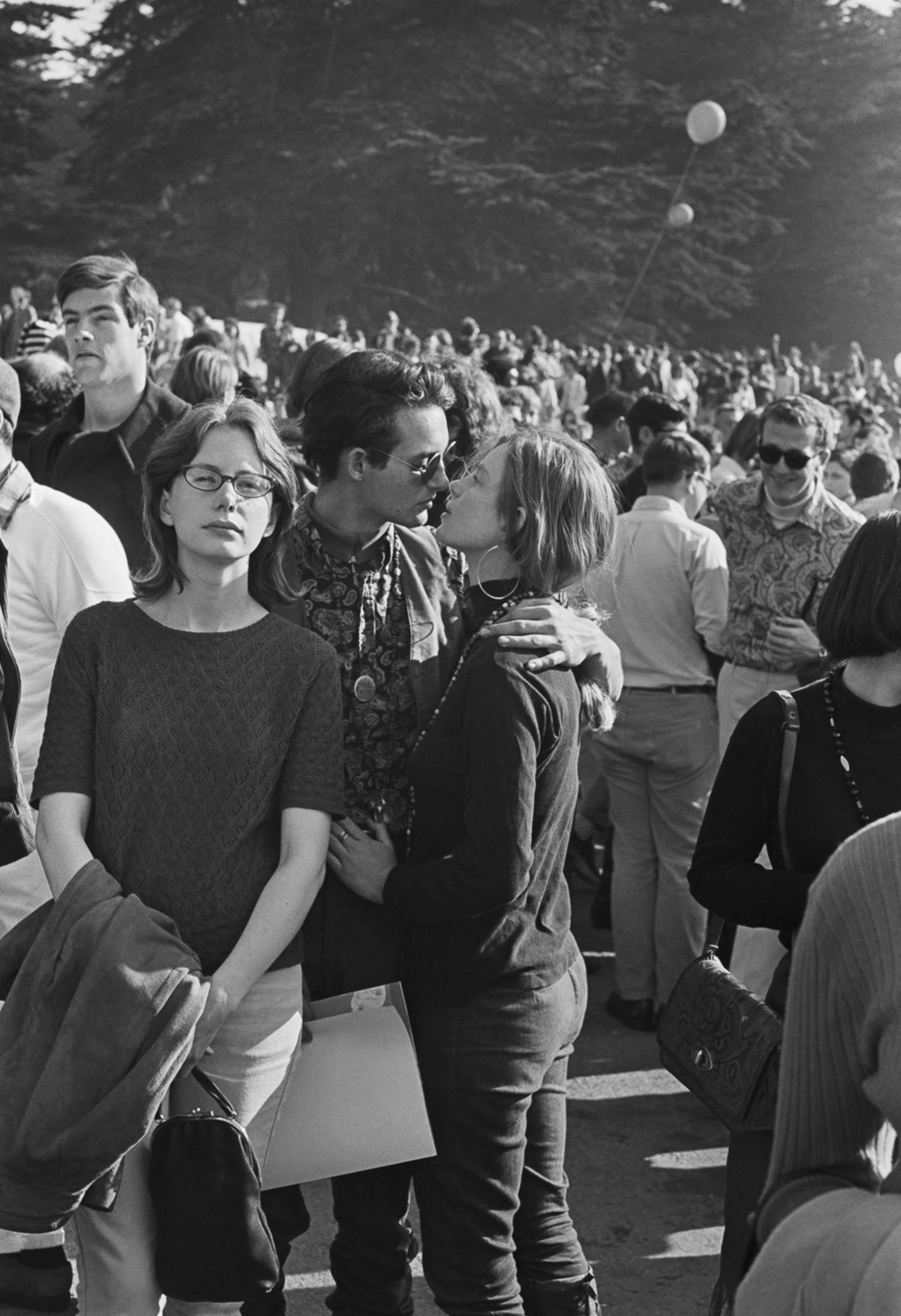 A black-and-white photo of a crowded outdoor event shows a couple kissing, surrounded by groups of people, with balloons floating in the background.