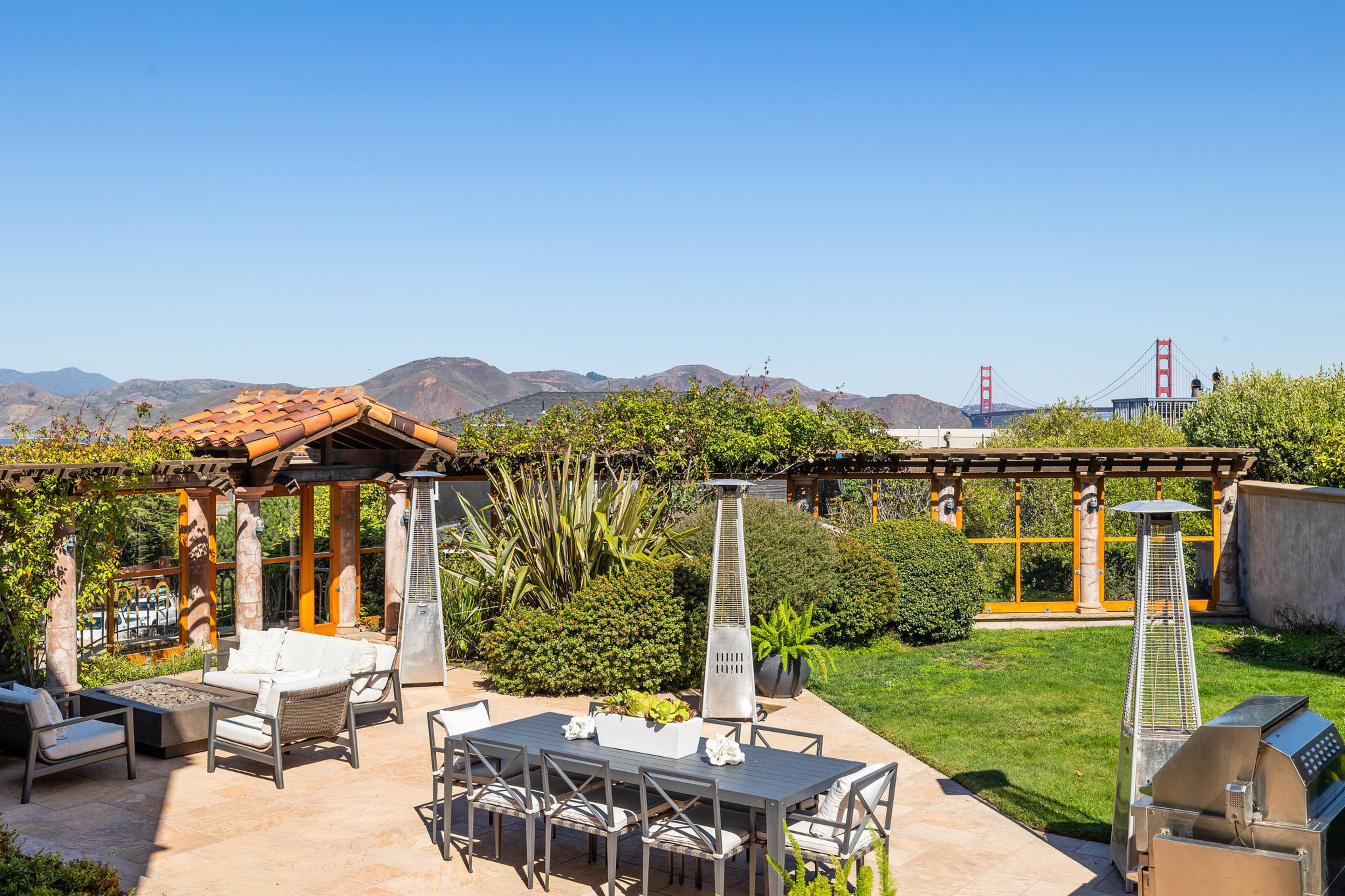 Outdoor patio features a dining table with eight chairs, lounge seating, two tall heaters, a barbecue grill, lush greenery, and the Golden Gate Bridge in the background.