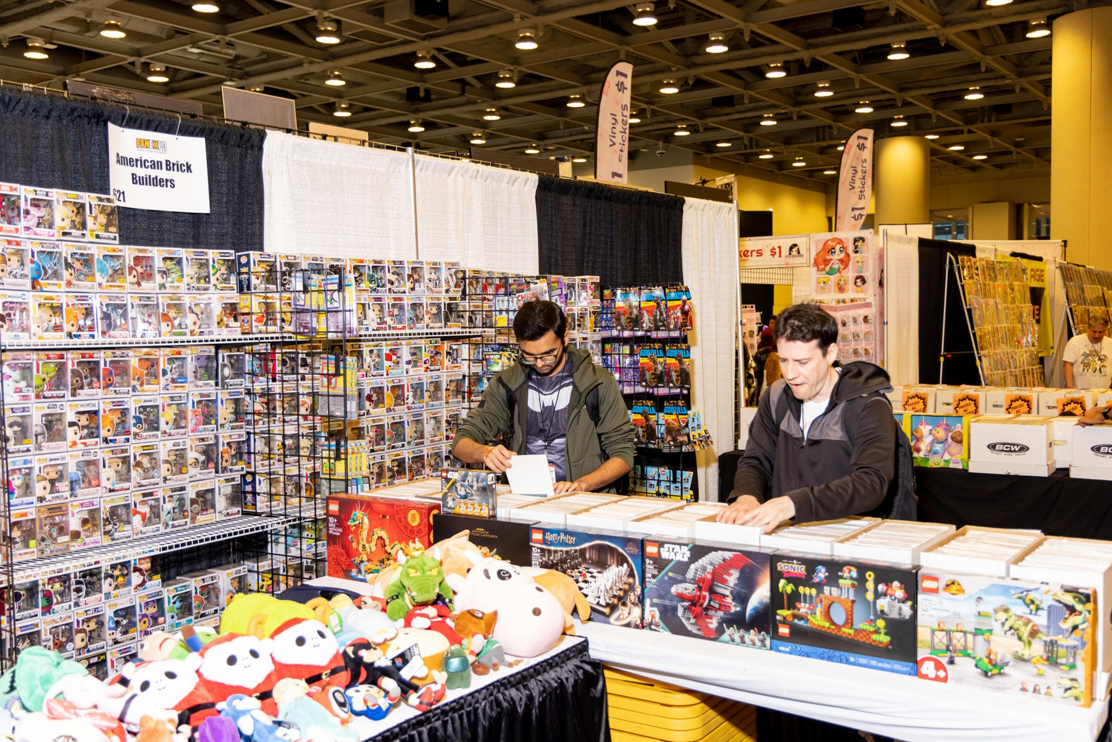 Two people are organizing merchandise at a convention booth. Shelves display collectibles like Funko Pop figures, while the table holds LEGO sets and plush toys.