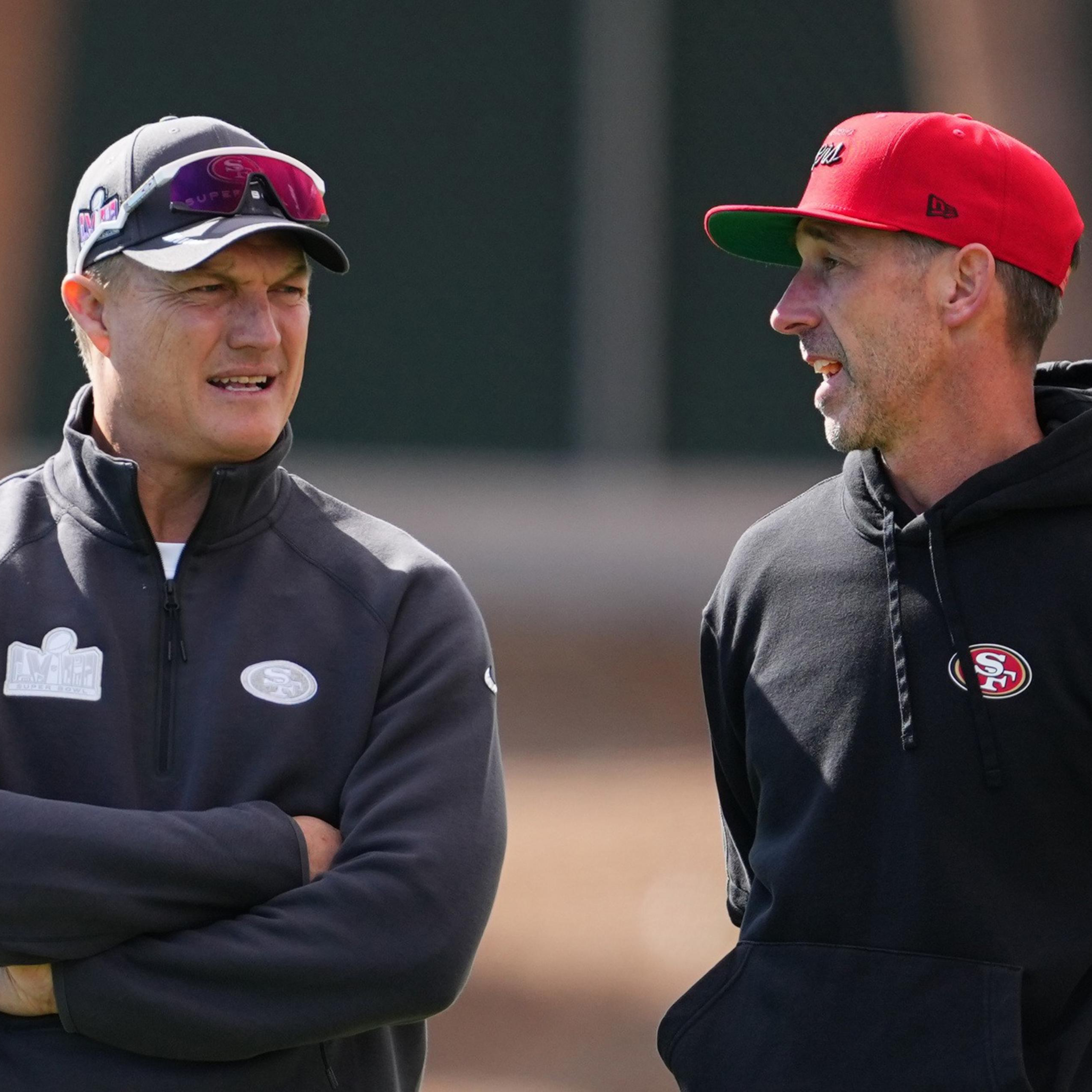 Two men are talking outdoors, wearing San Francisco 49ers gear. One has a gray jacket and sunglasses on his cap, while the other wears a red cap and black hoodie.