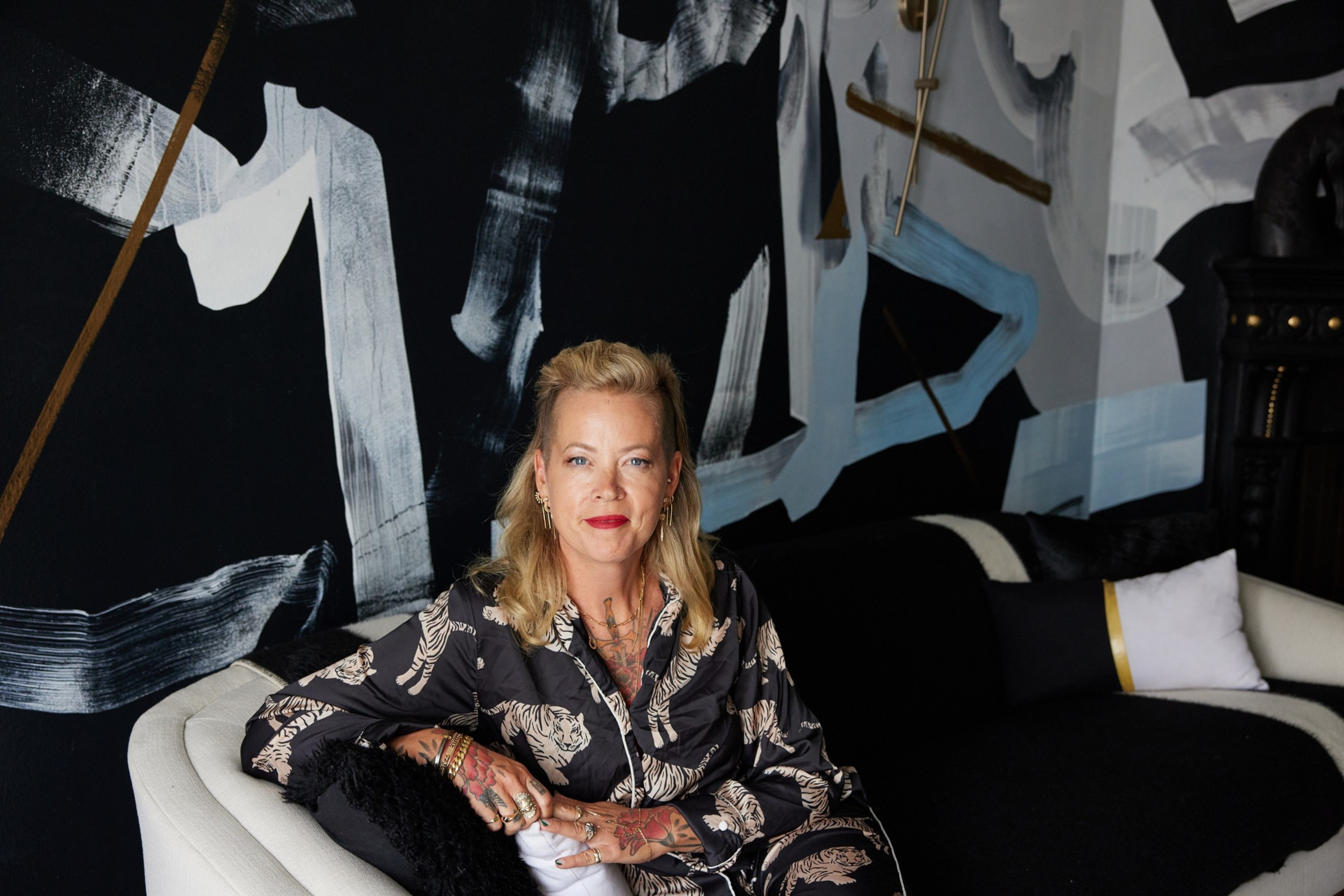 A blonde woman with tattoos sits on a white couch in front of a black and white abstract wall, wearing a patterned outfit and red lipstick.