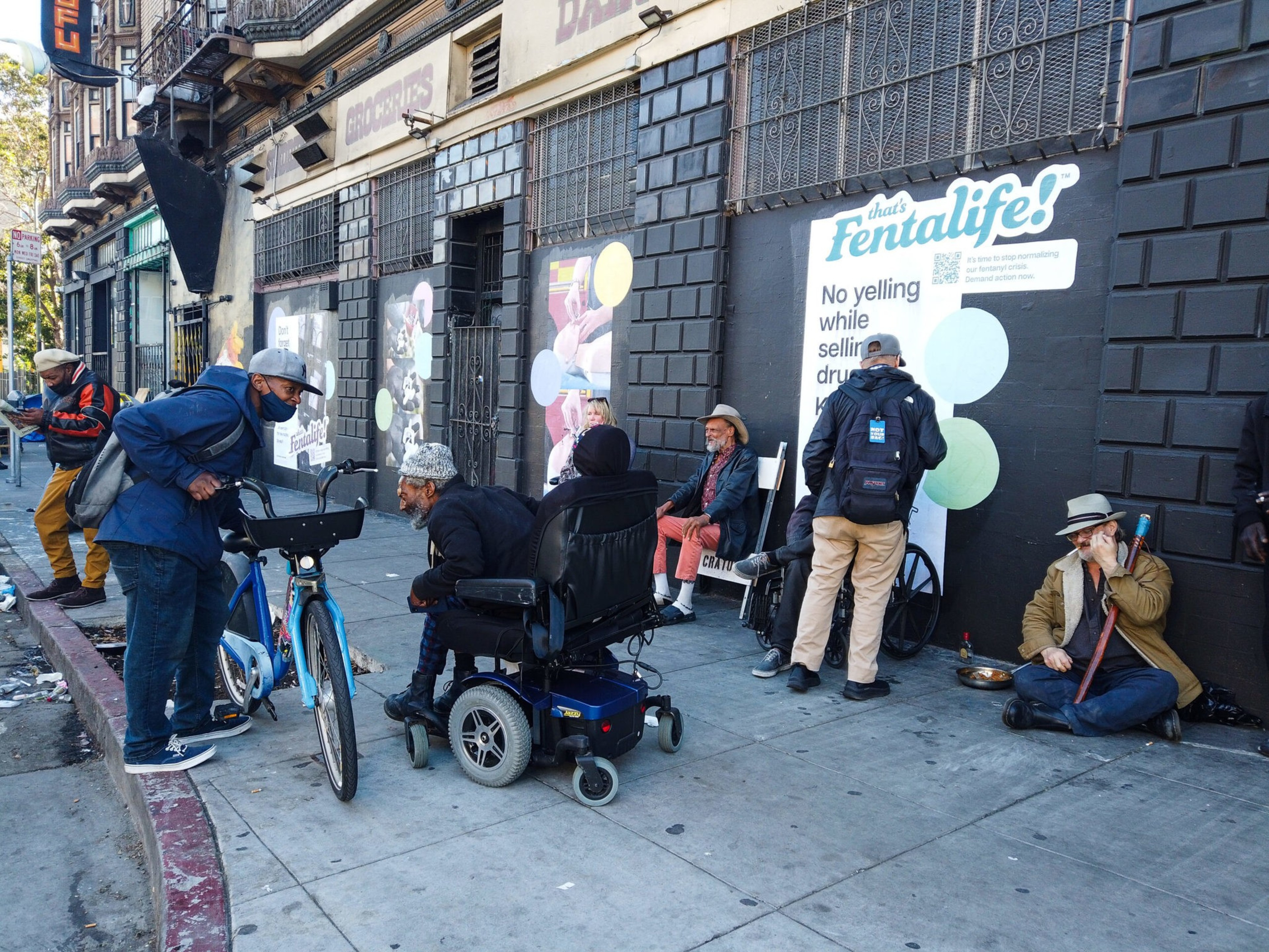 Several people gather on a city sidewalk near a building with a mural promoting "Fentalife," some sitting, one in a wheelchair, and one with a bike.