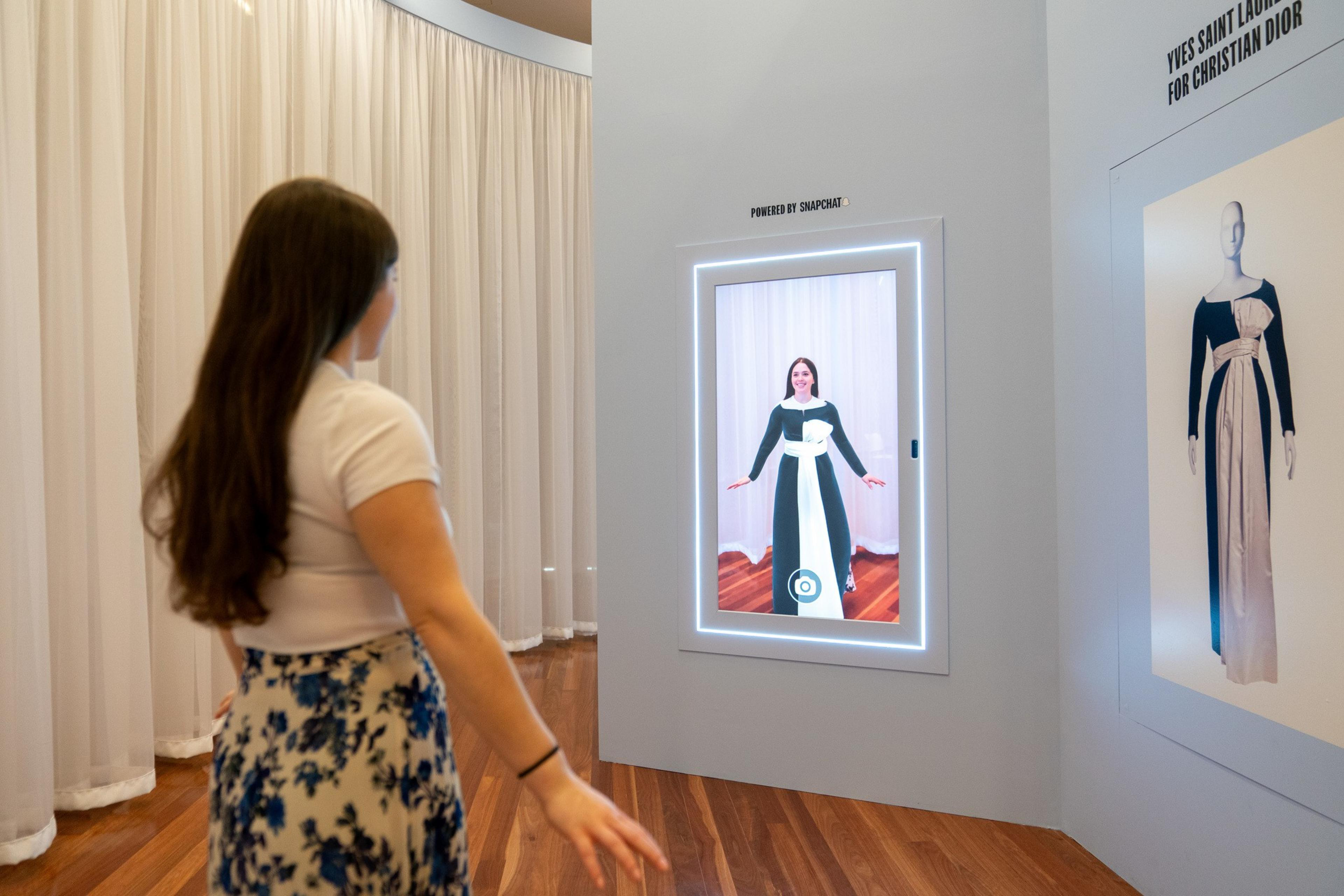 A woman stands in front of a screen in a black and white dress, with a different black and white dress reflected in the screen.