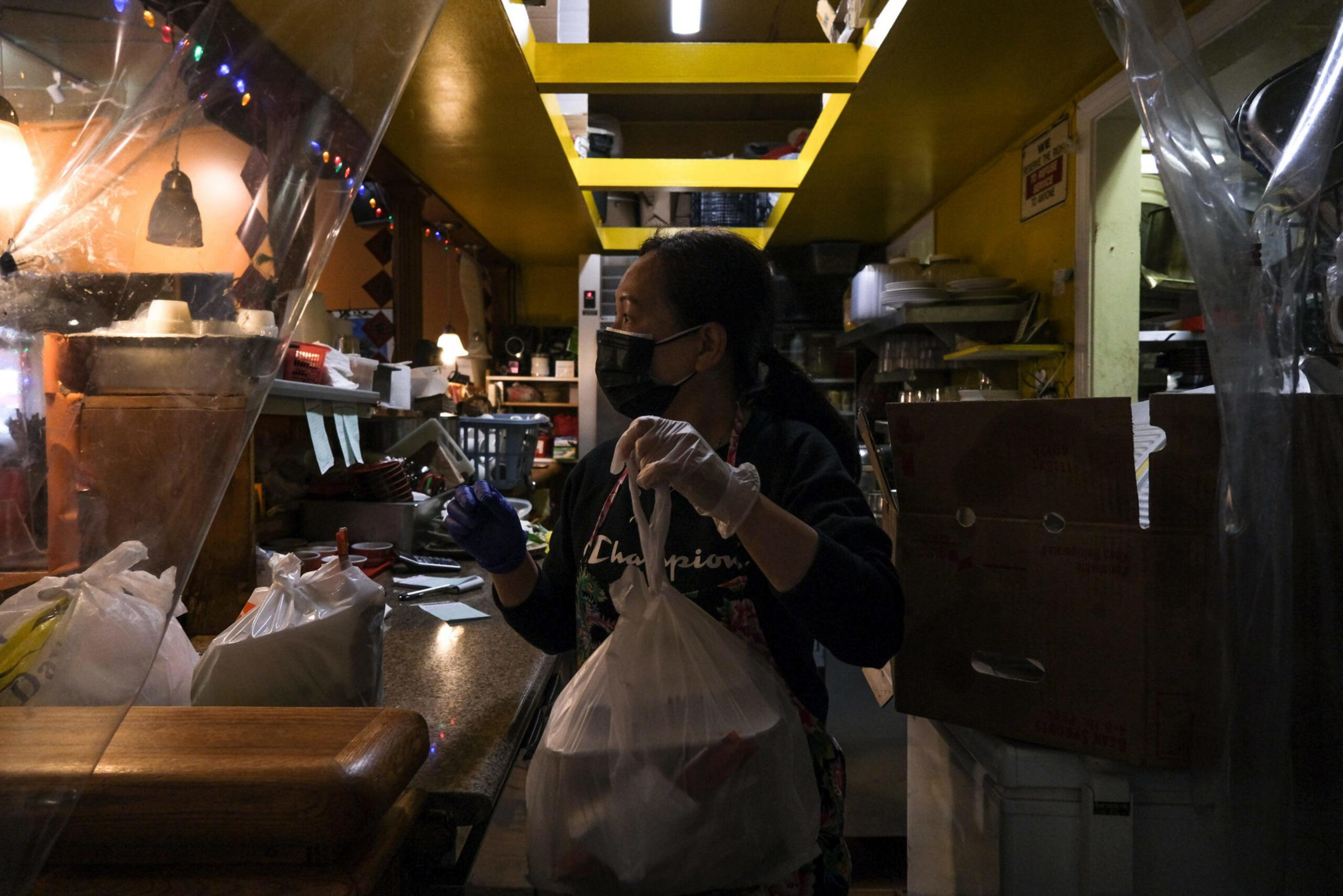 A person wearing a mask and gloves holds a plastic bag in a dimly lit restaurant or food service area, surrounded by boxes and shelves.