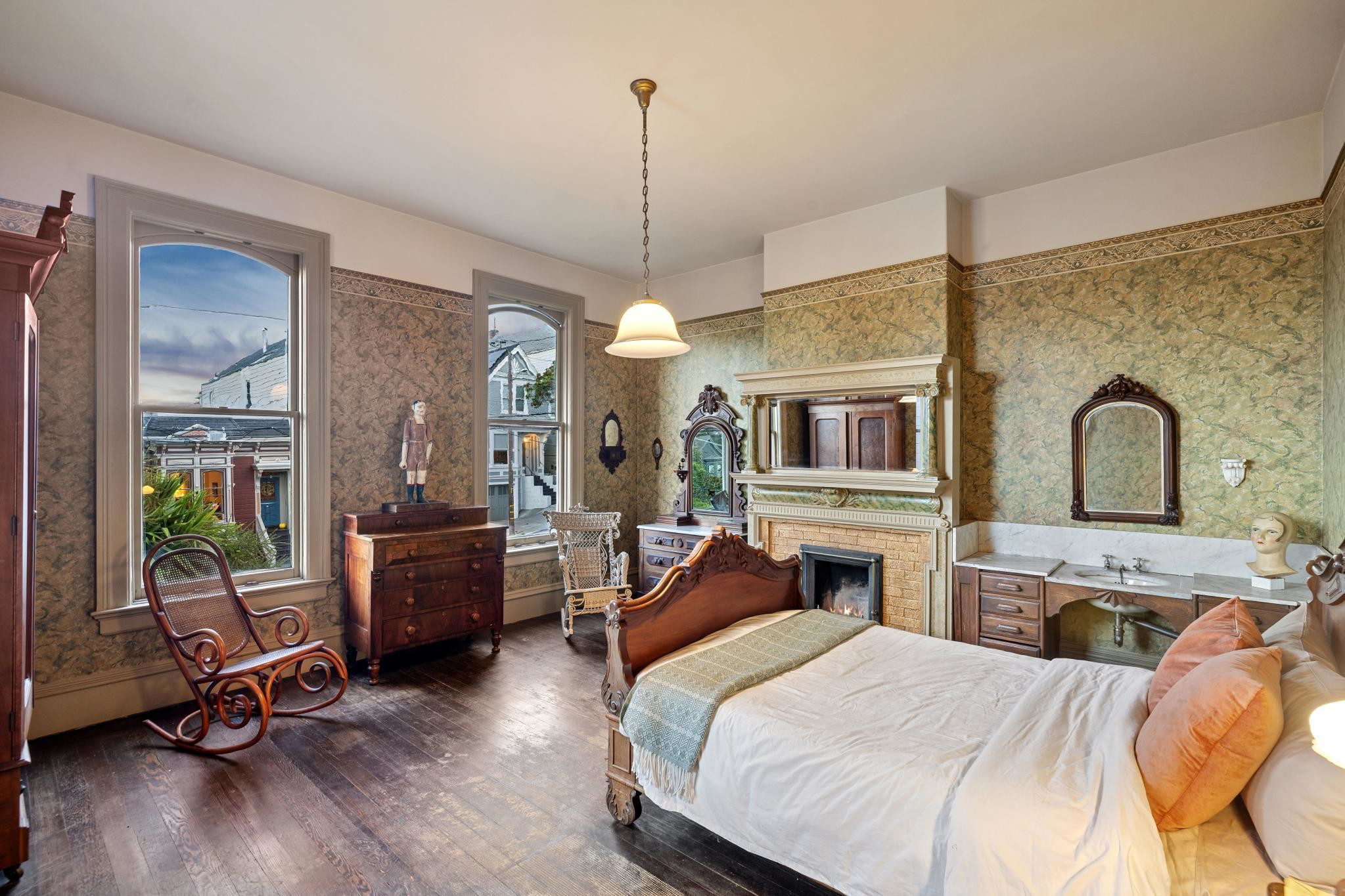 A vintage bedroom with wooden floor, carved bed, rocking chair, dresser, fireplace, twin windows, and a vanity with a mirror and sink.