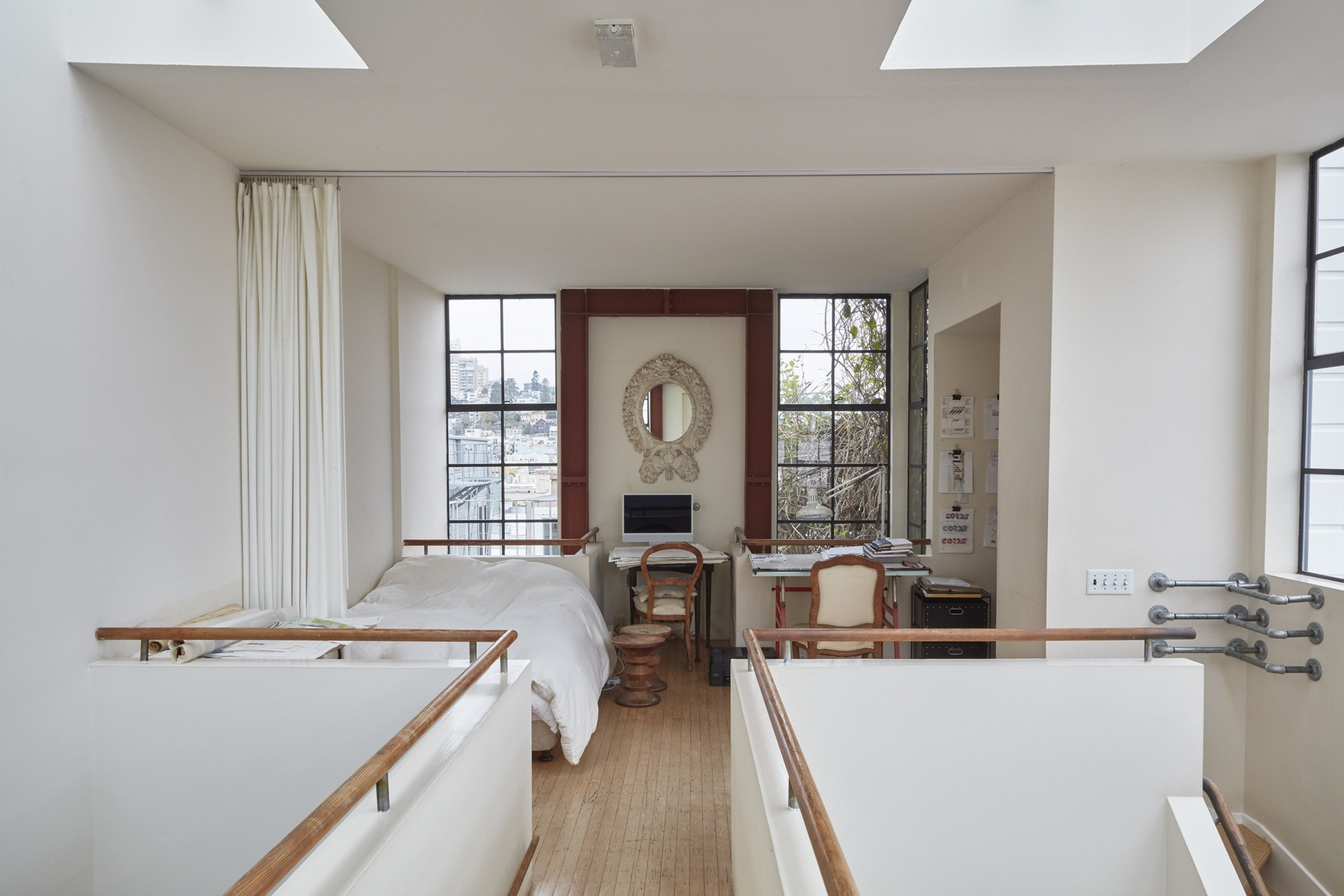 A cozy bedroom with a double bed and a small desk overlooks large windows. The room has a minimalistic design with light colors and a wooden floor.