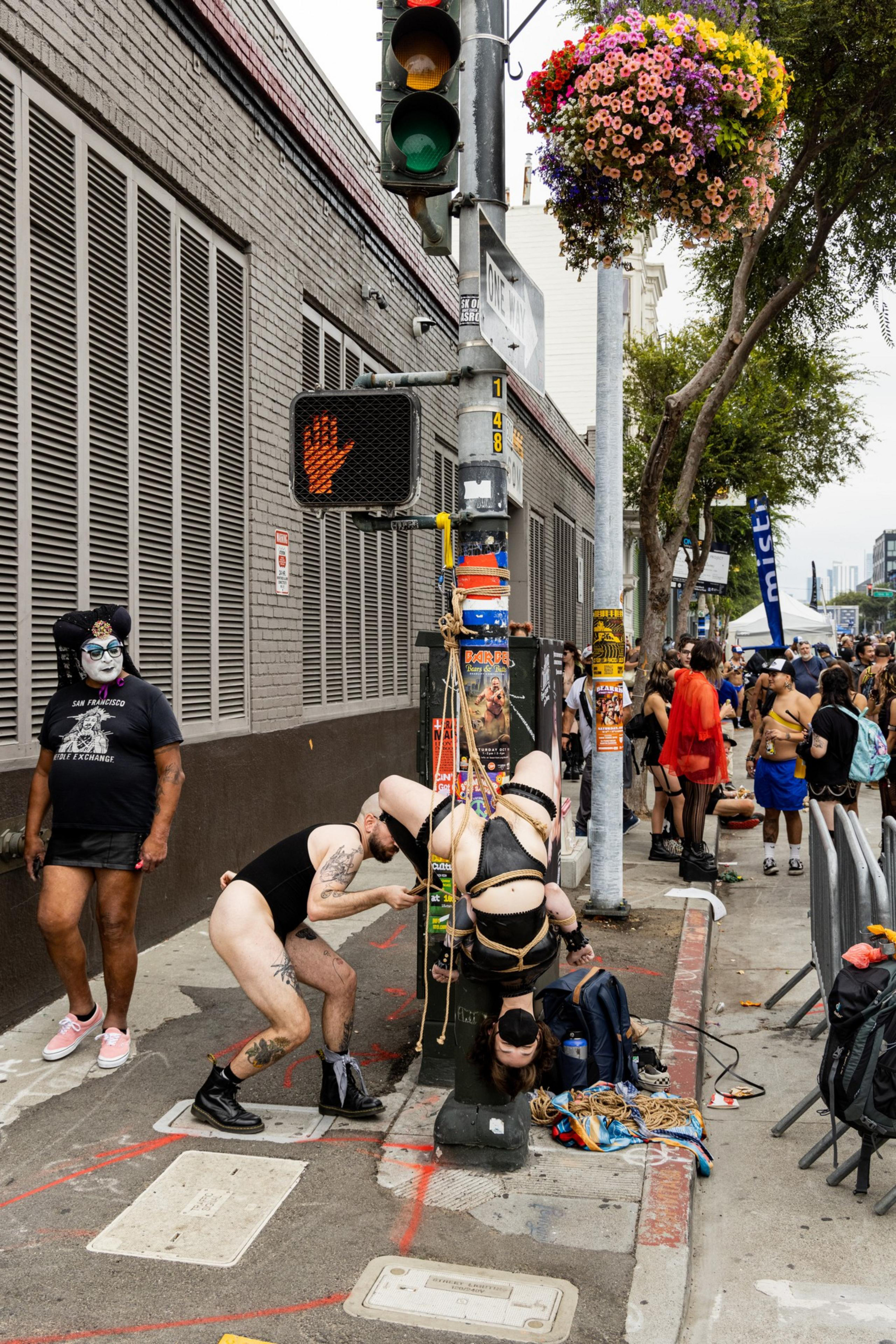 A person is suspended upside down by ropes attached to a pole while another man adjusts the ropes, as a crowd gathers on the sidewalk nearby.