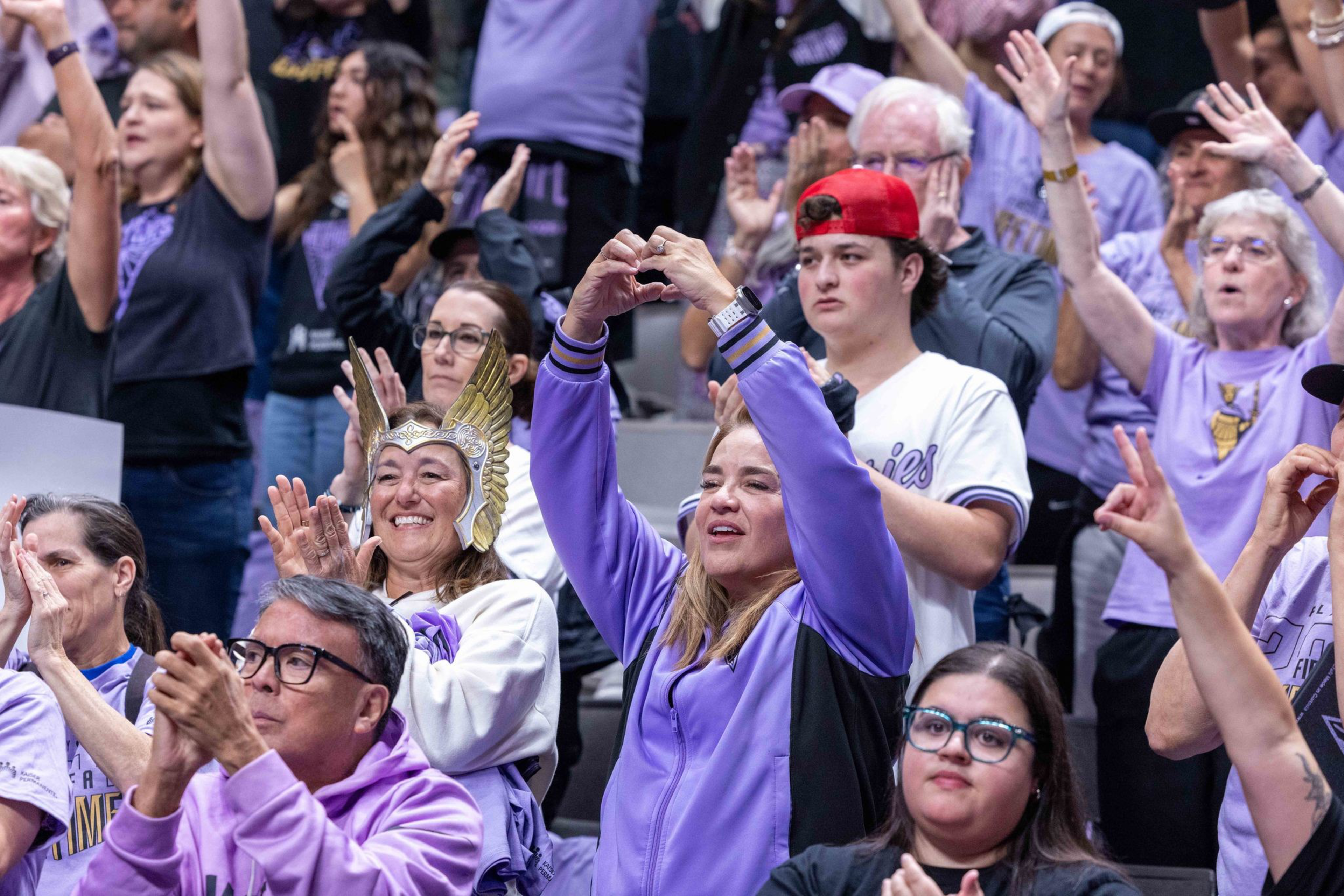 A crowd of fans wearing purple cheer and applaud, with one woman making a heart shape with her hands and another smiling with a winged helmet.