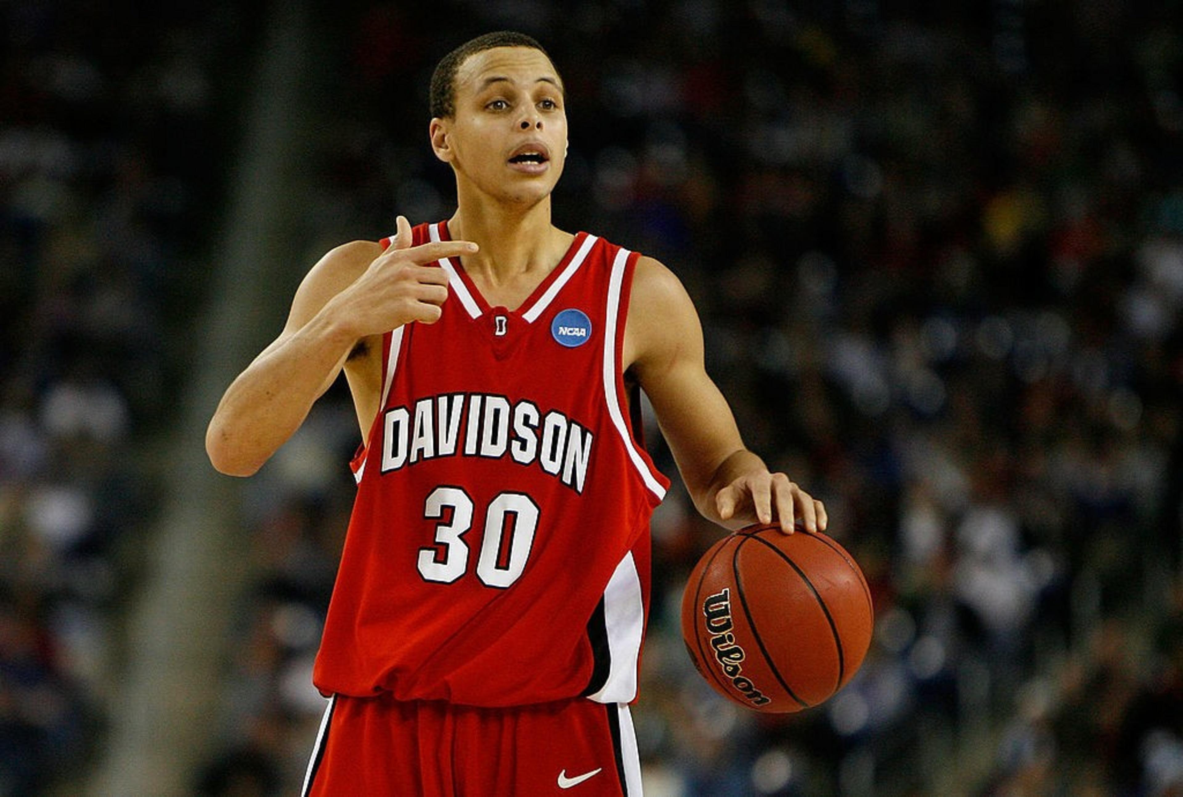A basketball player in a red Davidson jersey with the number 30 holds the ball, gesturing with his left hand. The background shows a blurred audience.