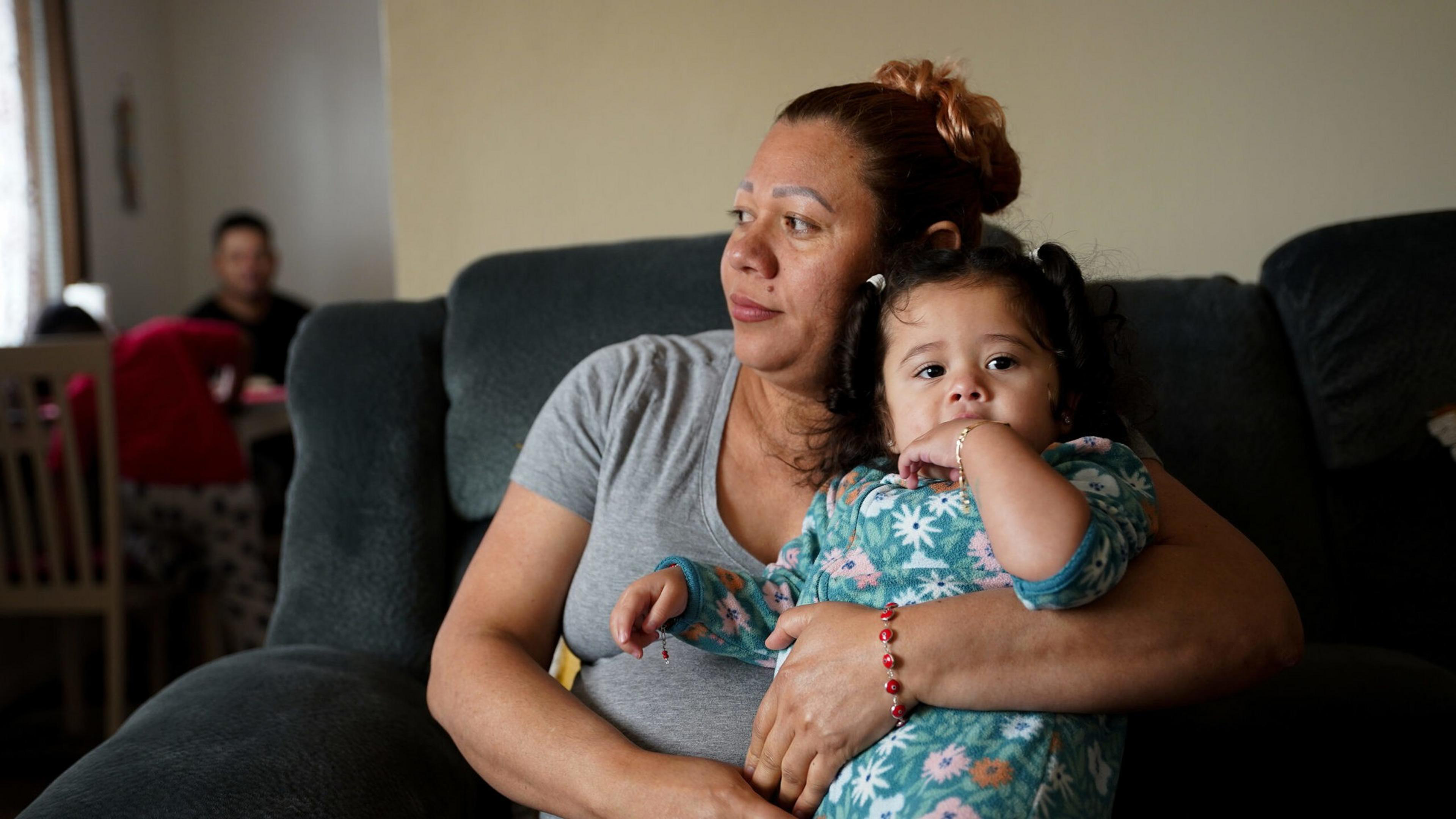 A woman with red hair tied up holds a young child in floral pajamas while sitting on a dark couch, with a person sitting in the background.