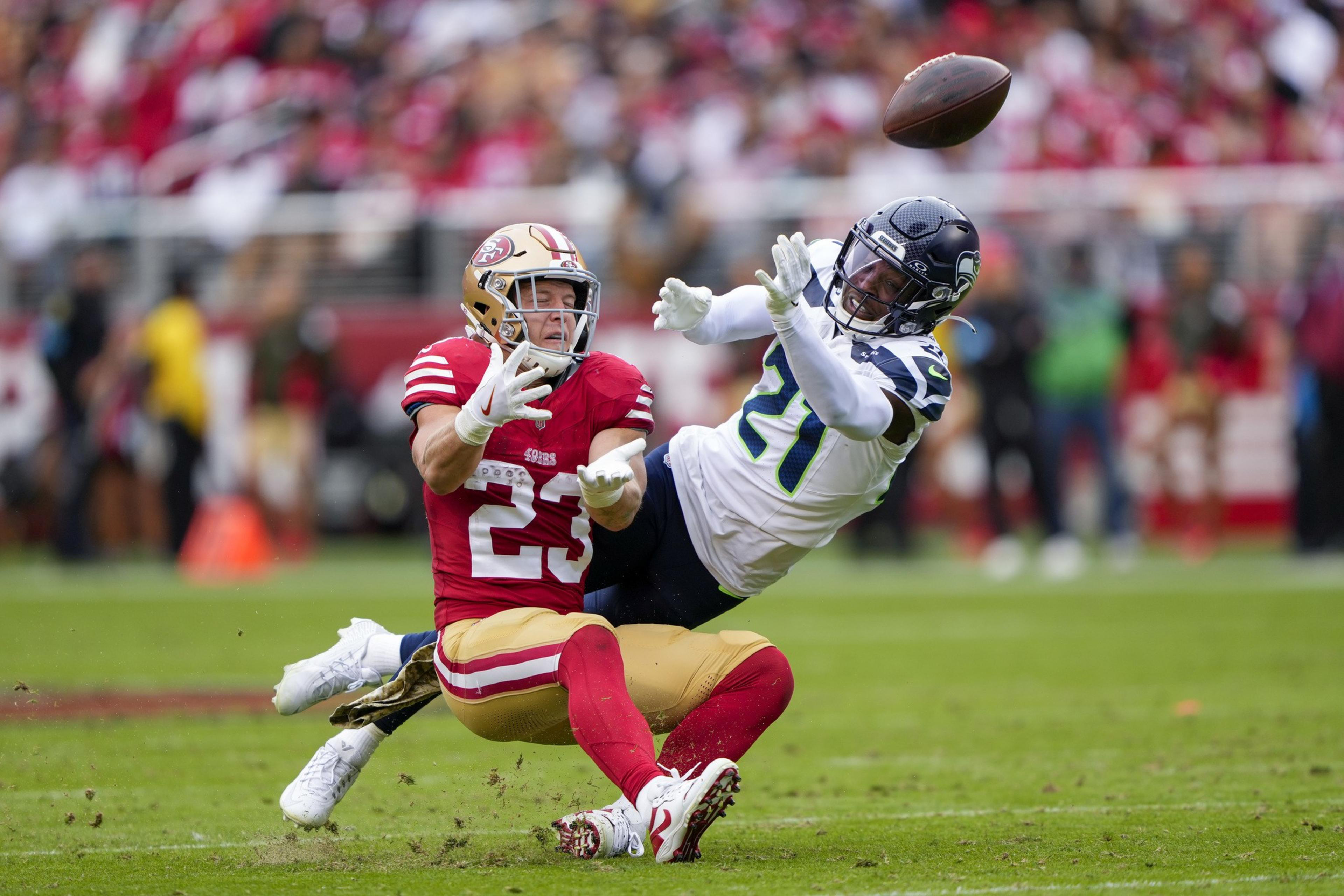 Two football players are in action; one in a red uniform is falling as he reaches for the ball, while the other in white tries to intercept it. The background is blurred.