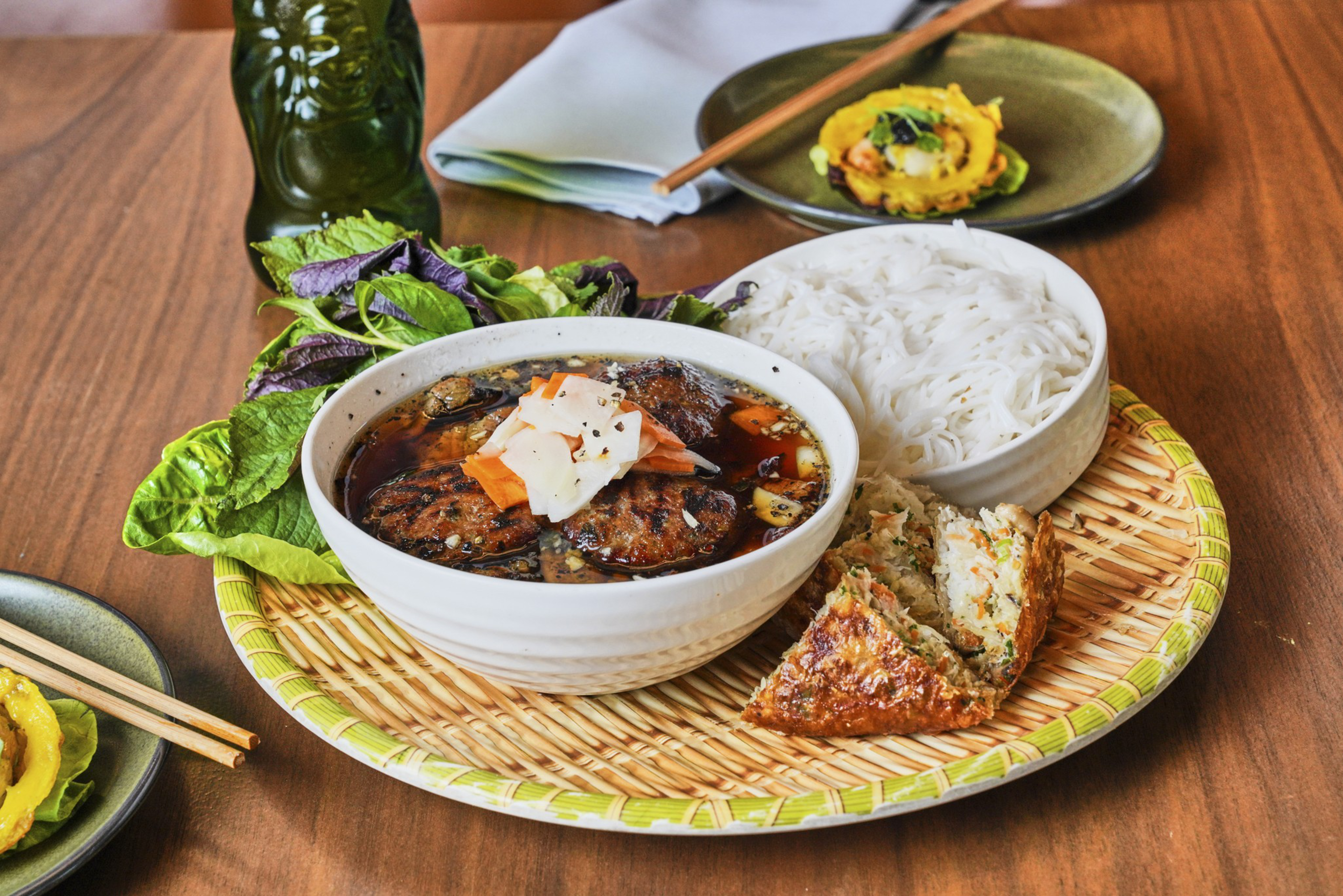 A platter displays a bowl of broth with meat patties topped with pickled veggies, rice noodles, fresh greens, and a fried savory pastry, set on a wood table.