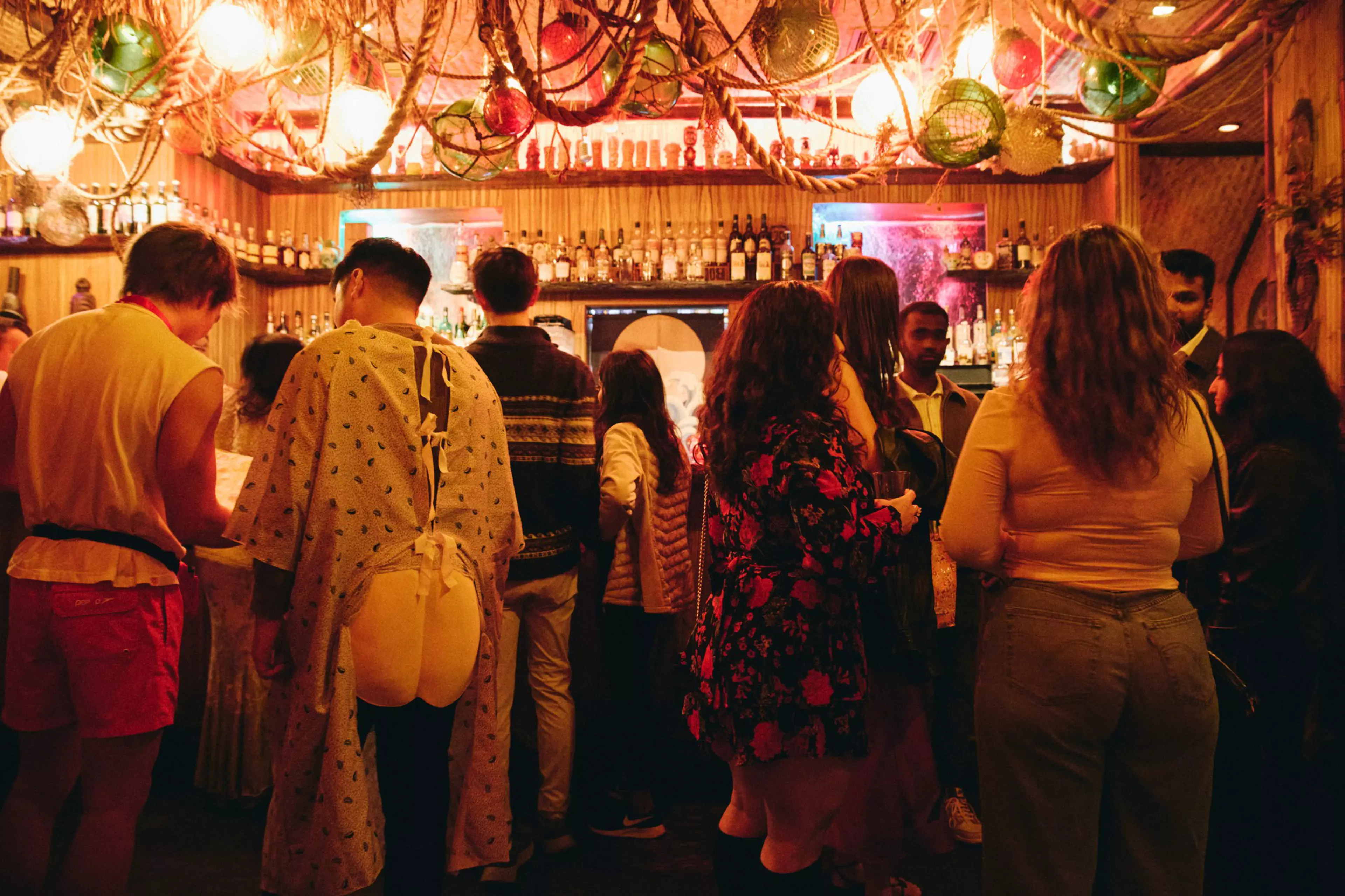 A group of people stands at a bar under warm, hanging lights with ropes and colorful glass floats decorating the ceiling. One person wears a hospital gown.