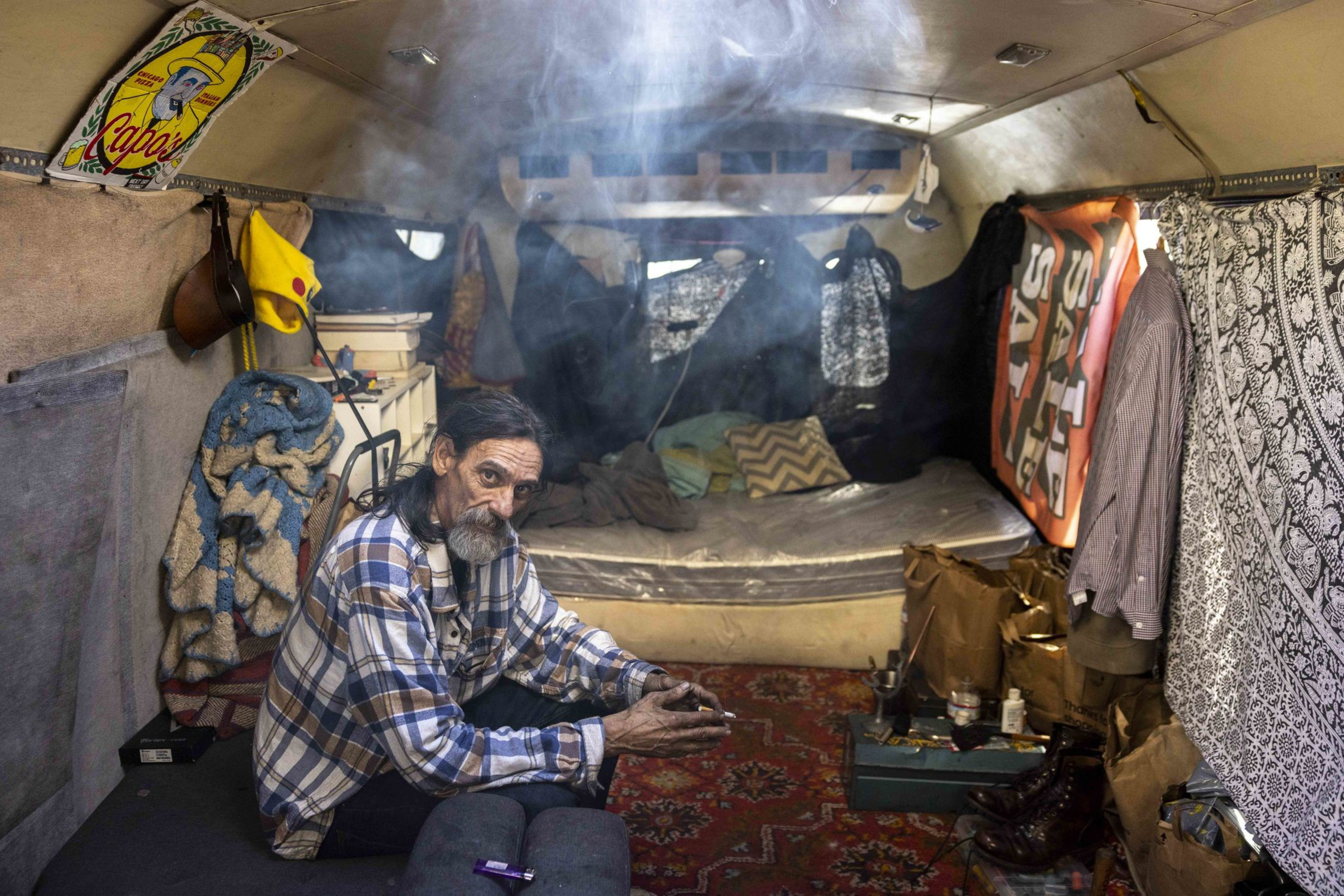 A man with a plaid shirt sits inside a cluttered camper with hanging clothes and blankets. Soft light filters through, creating a smoky, cozy atmosphere.