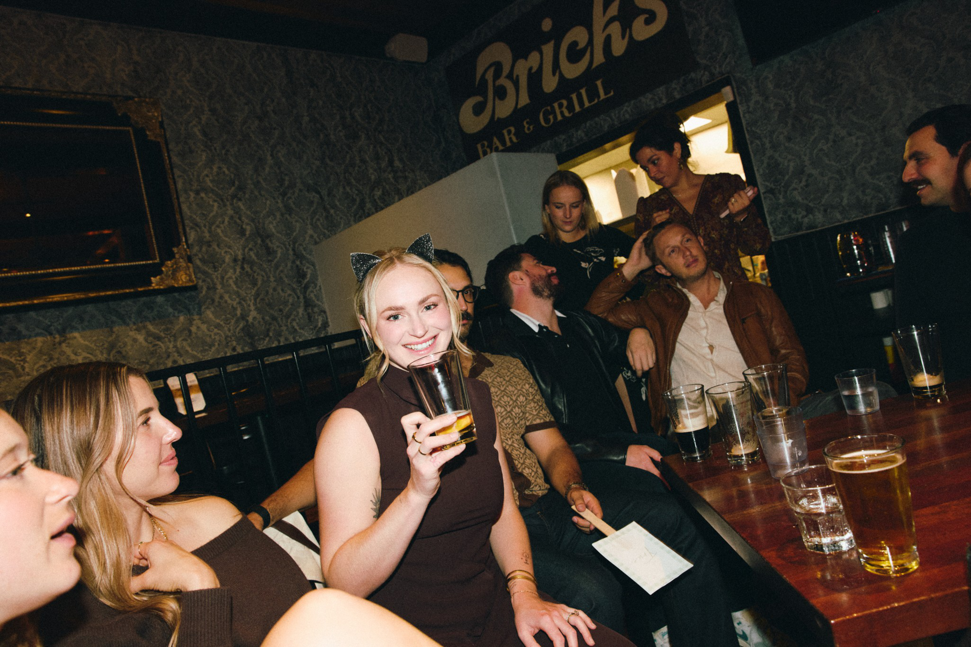 A group of friends sit closely at a bar table with drinks, smiling and chatting, with one woman wearing cat ears and holding a glass.