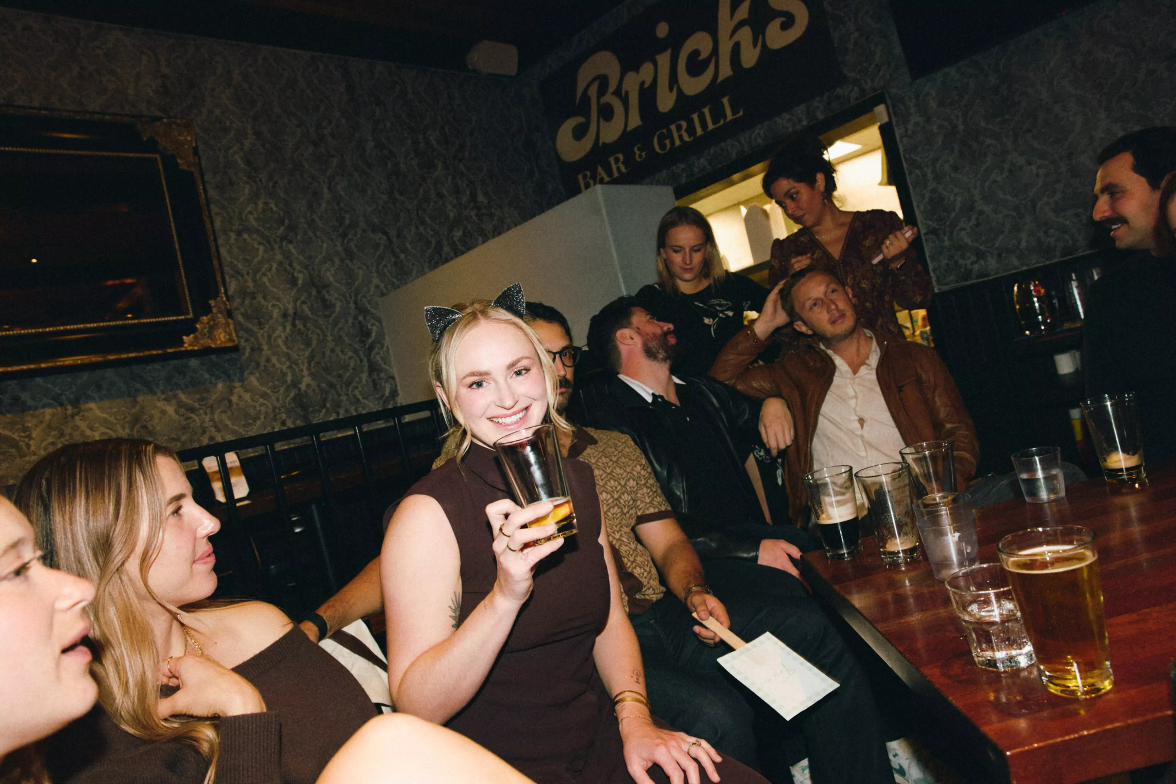 A group of friends sit closely at a bar table with drinks, smiling and chatting, with one woman wearing cat ears and holding a glass.