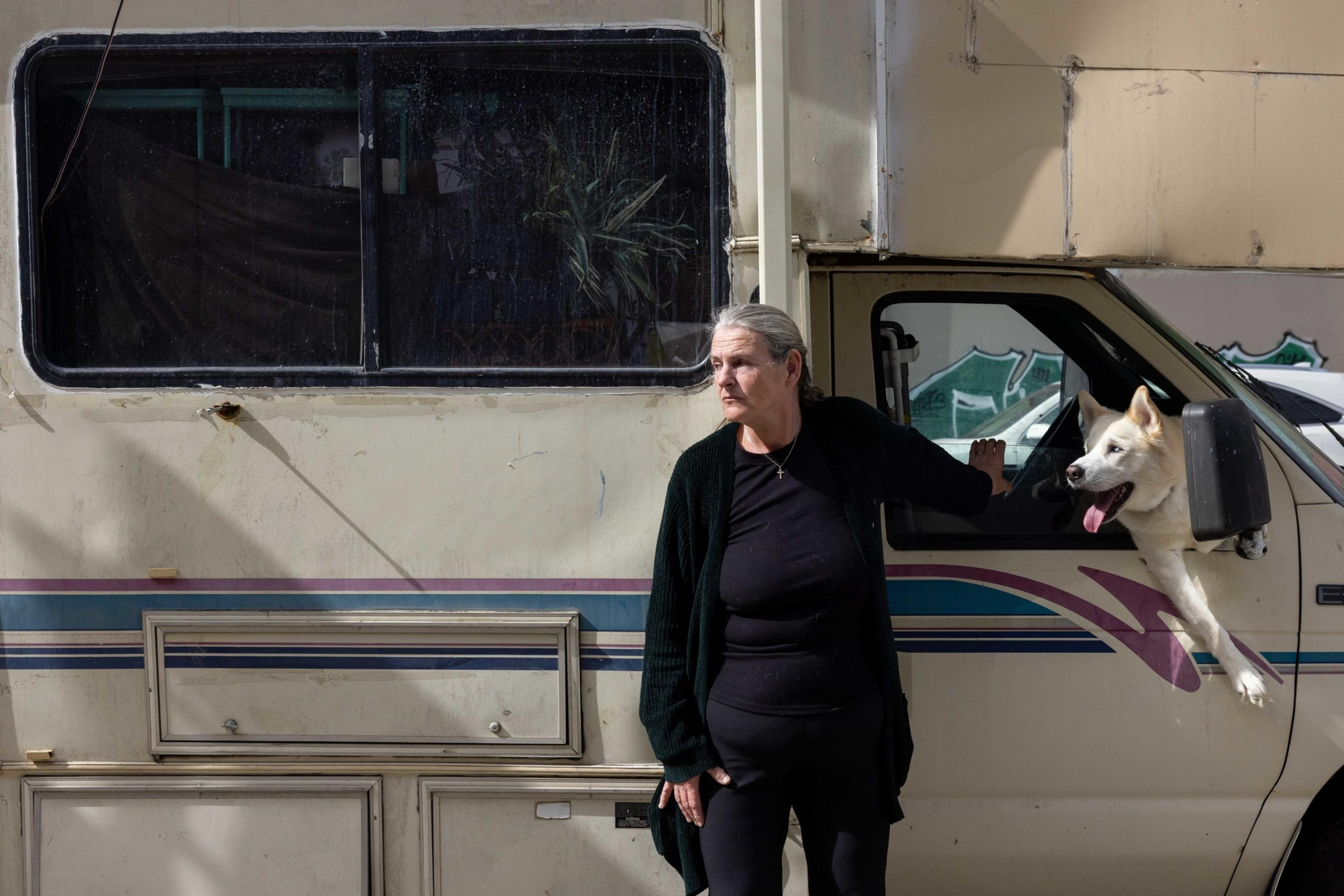 A woman in black stands leaning against an RV, with a white dog sticking its head and paws out of the driver's window. The RV is parked on a street.