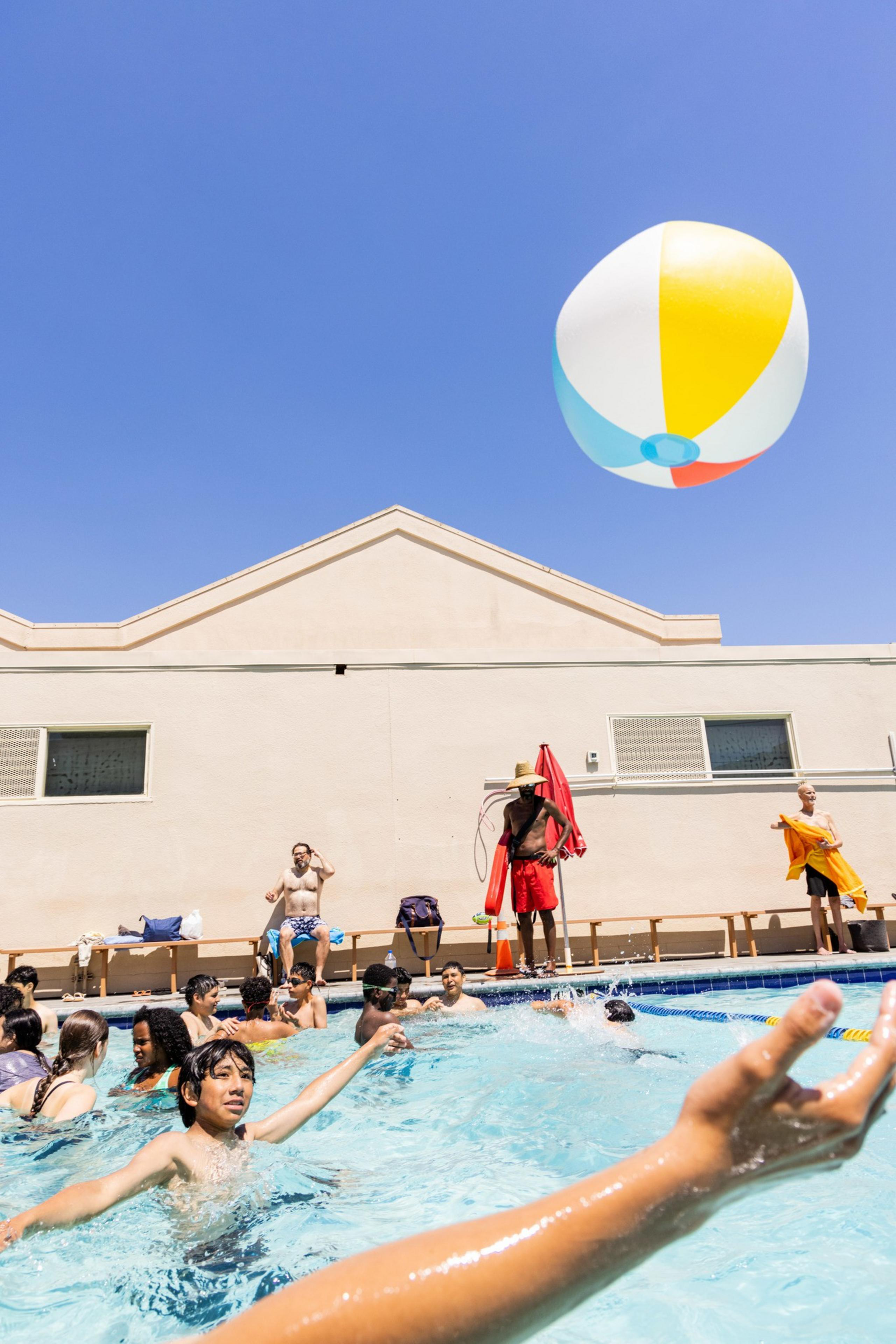 Children are playing with a large beach ball in a swimming pool under a blue sky, while two lifeguards watch from the poolside. A building is visible in the background.