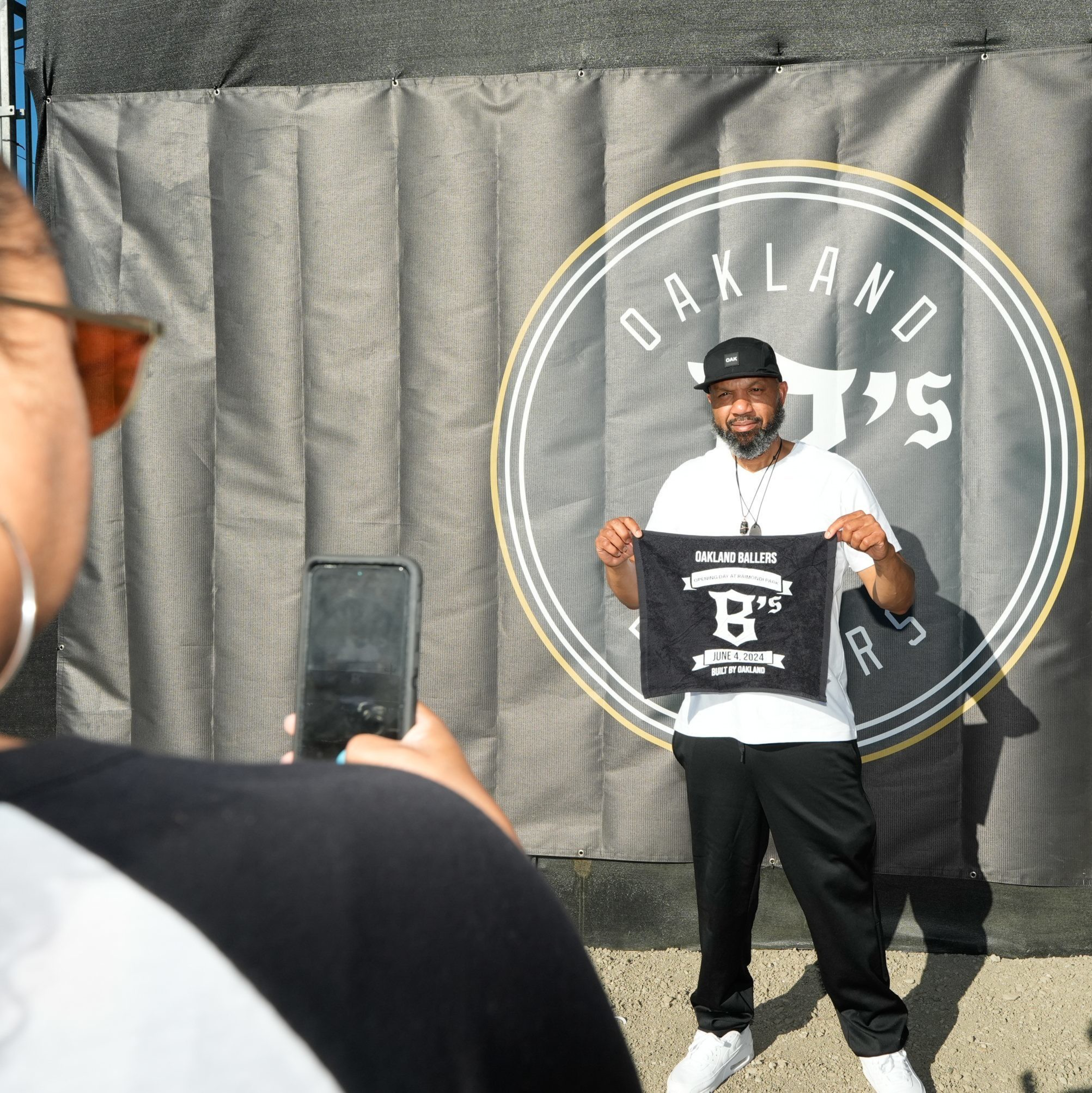 A man stands in front of a backdrop with the text &quot;Oakland B's&quot; while holding a sign and posing for a woman taking his photo with a phone.