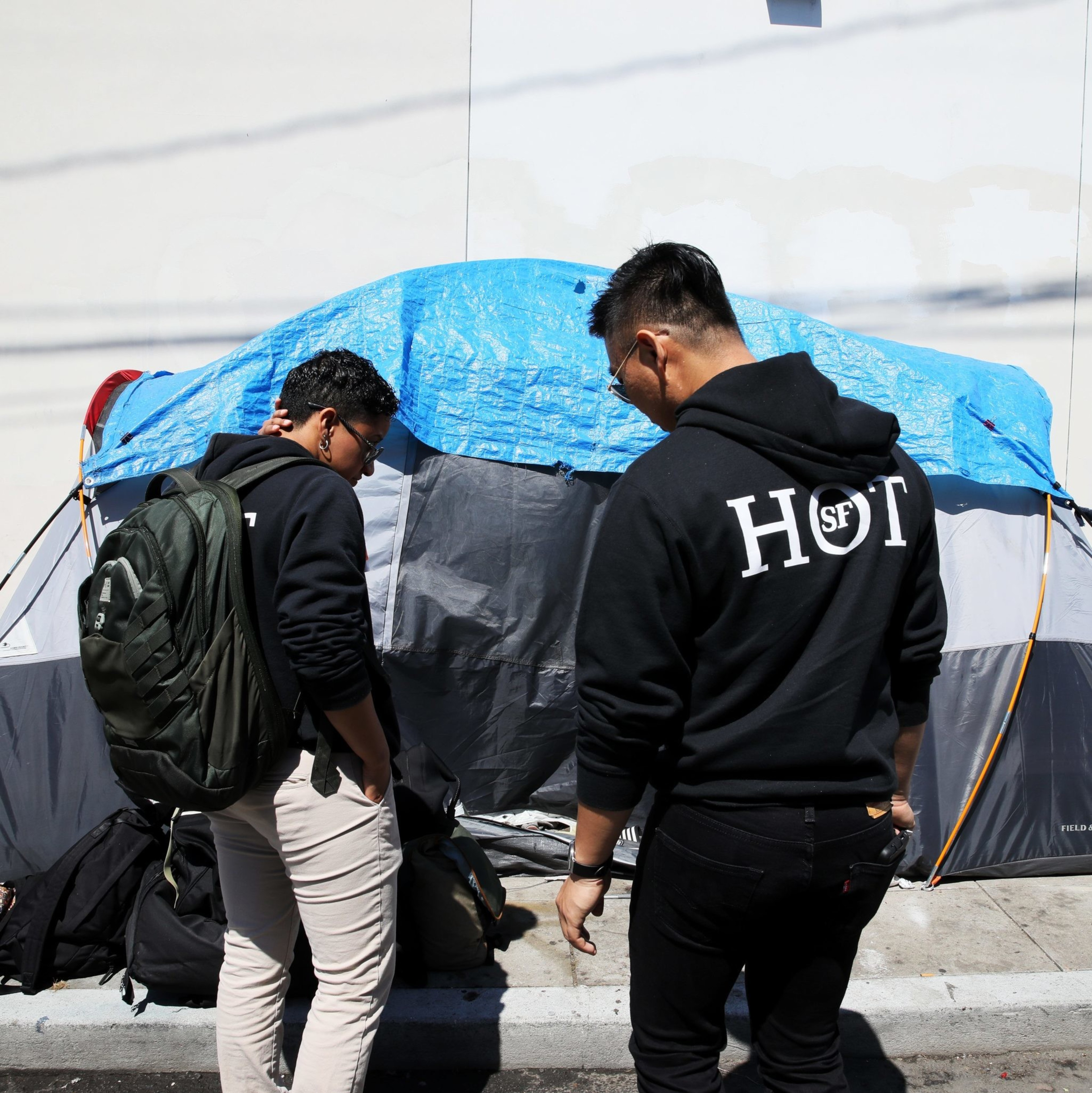 Two individuals, wearing &quot;HOT SF&quot; hoodies and one with a backpack, stand outside a makeshift tent covered with a blue tarp on a sidewalk, next to a shopping cart.