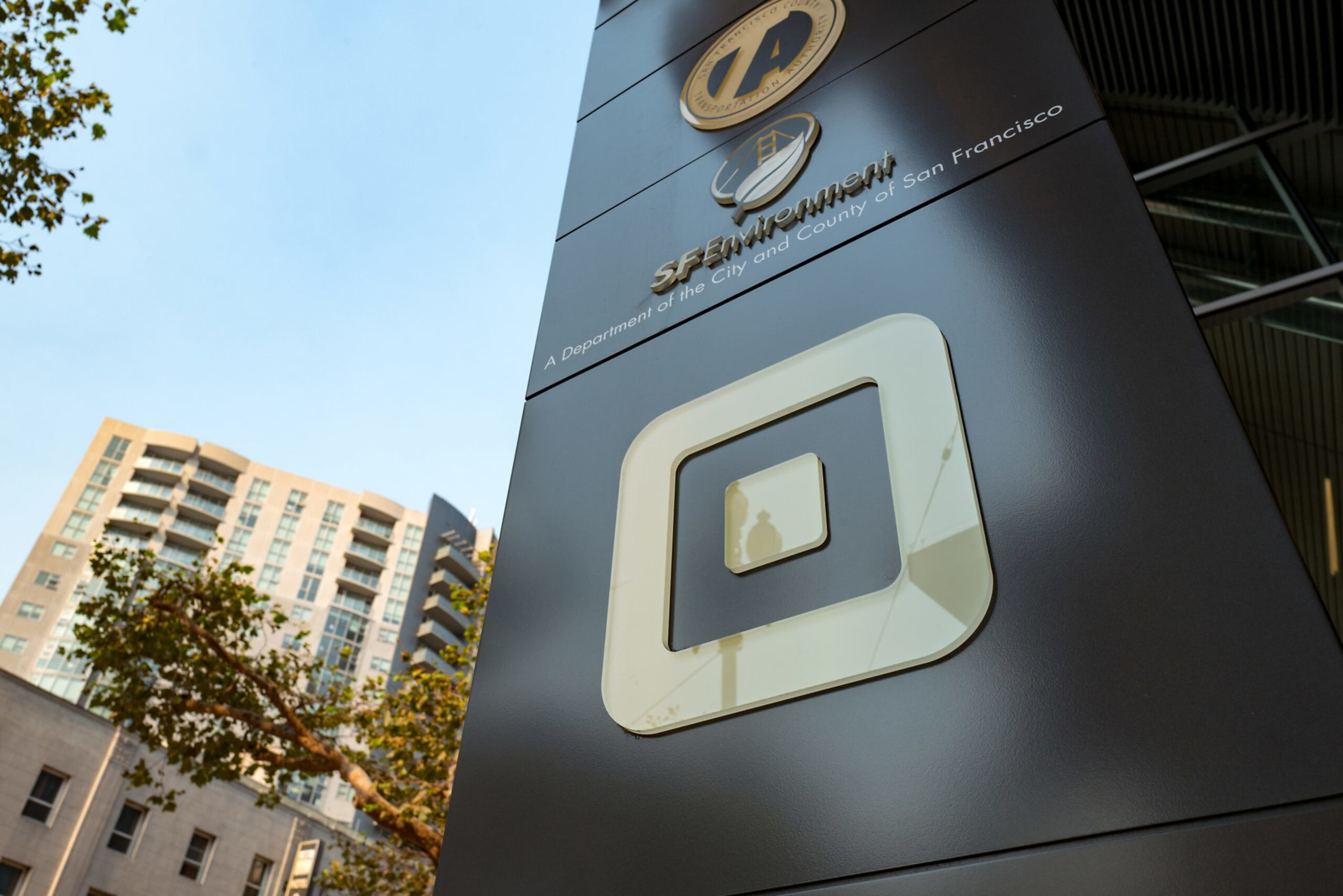 A black pillar displays the SF Environment logo and text reading "A Department of the City and County of San Francisco" against a backdrop of buildings and trees.