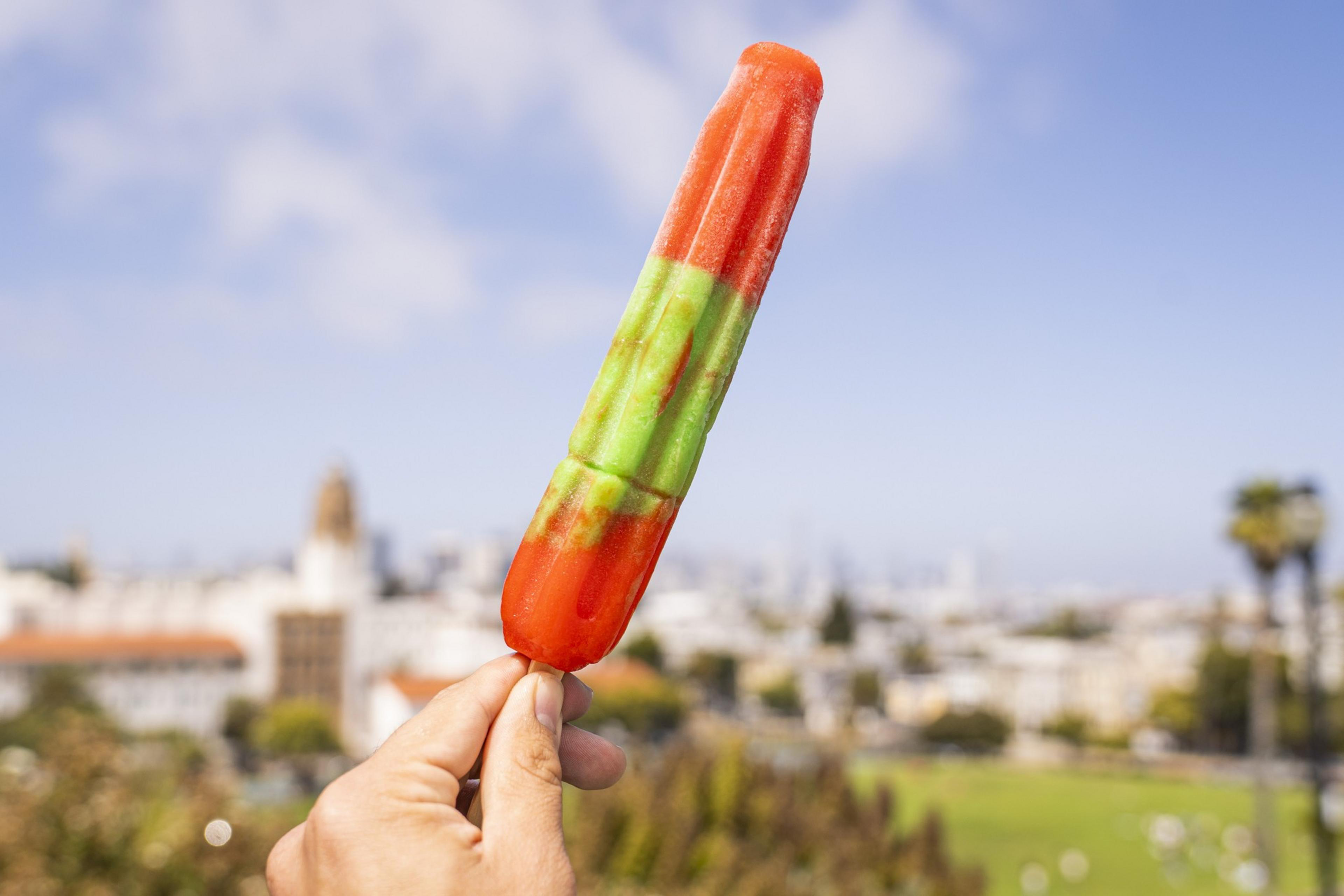 A hand holds a colorful popsicle with red, green, and orange sections against a blurred cityscape backdrop and blue sky.