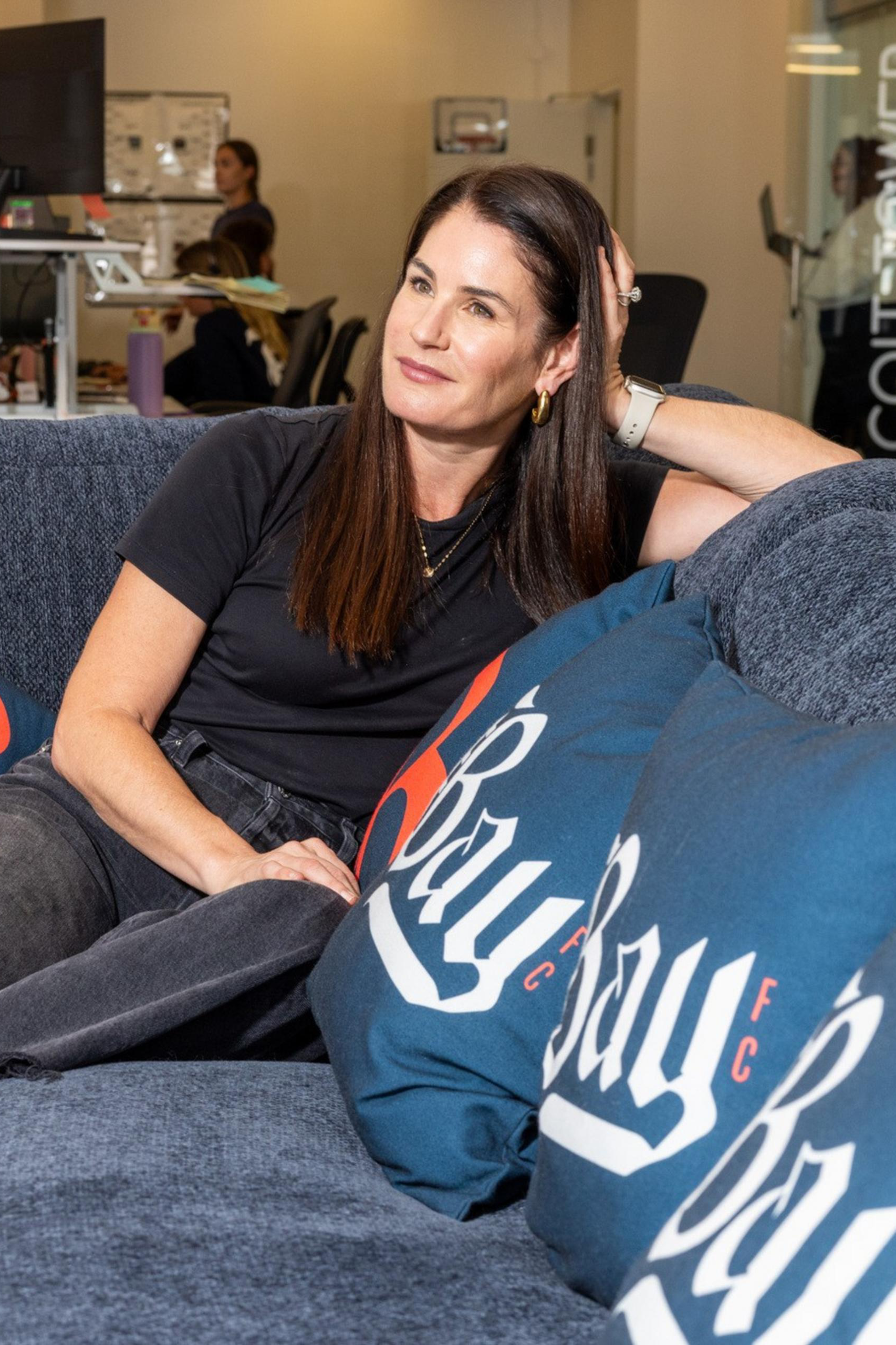 A woman with long dark hair sits relaxed on a blue couch adorned with decorative pillows, in a modern office setting with colleagues working in the background.