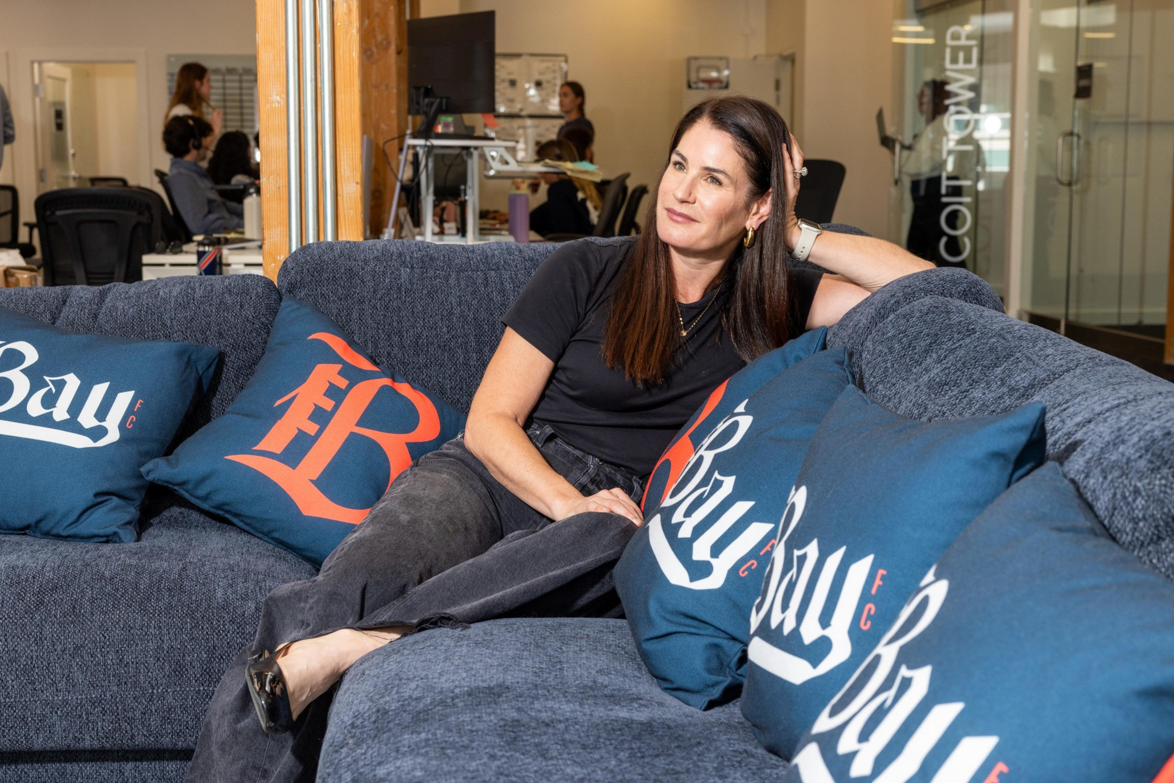 A woman with long dark hair sits relaxed on a blue couch adorned with decorative pillows, in a modern office setting with colleagues working in the background.