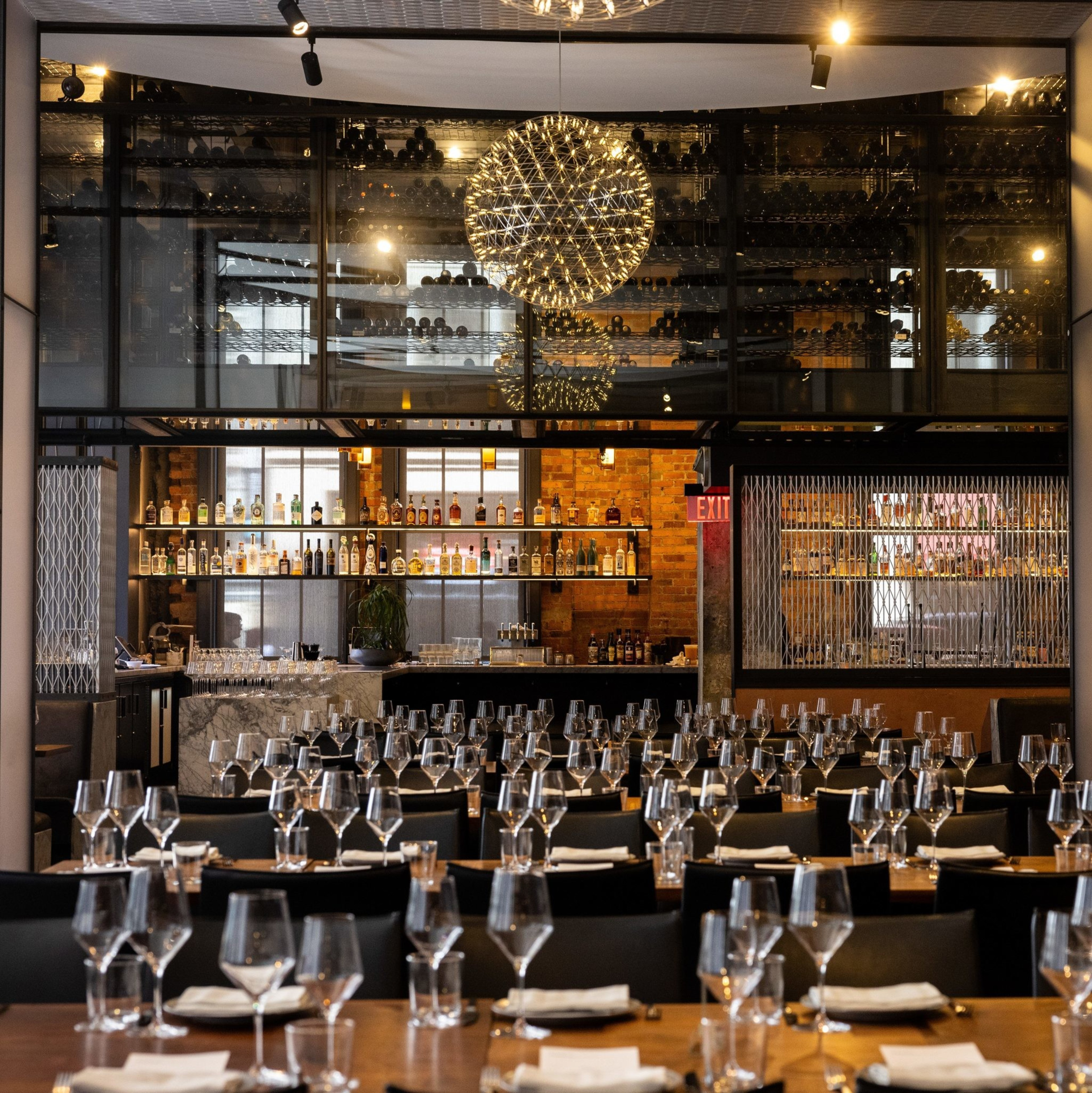 A stylish restaurant features elegantly set tables with neatly arranged glassware and napkins, a well-stocked bar in the background, and a shimmering spherical chandelier.