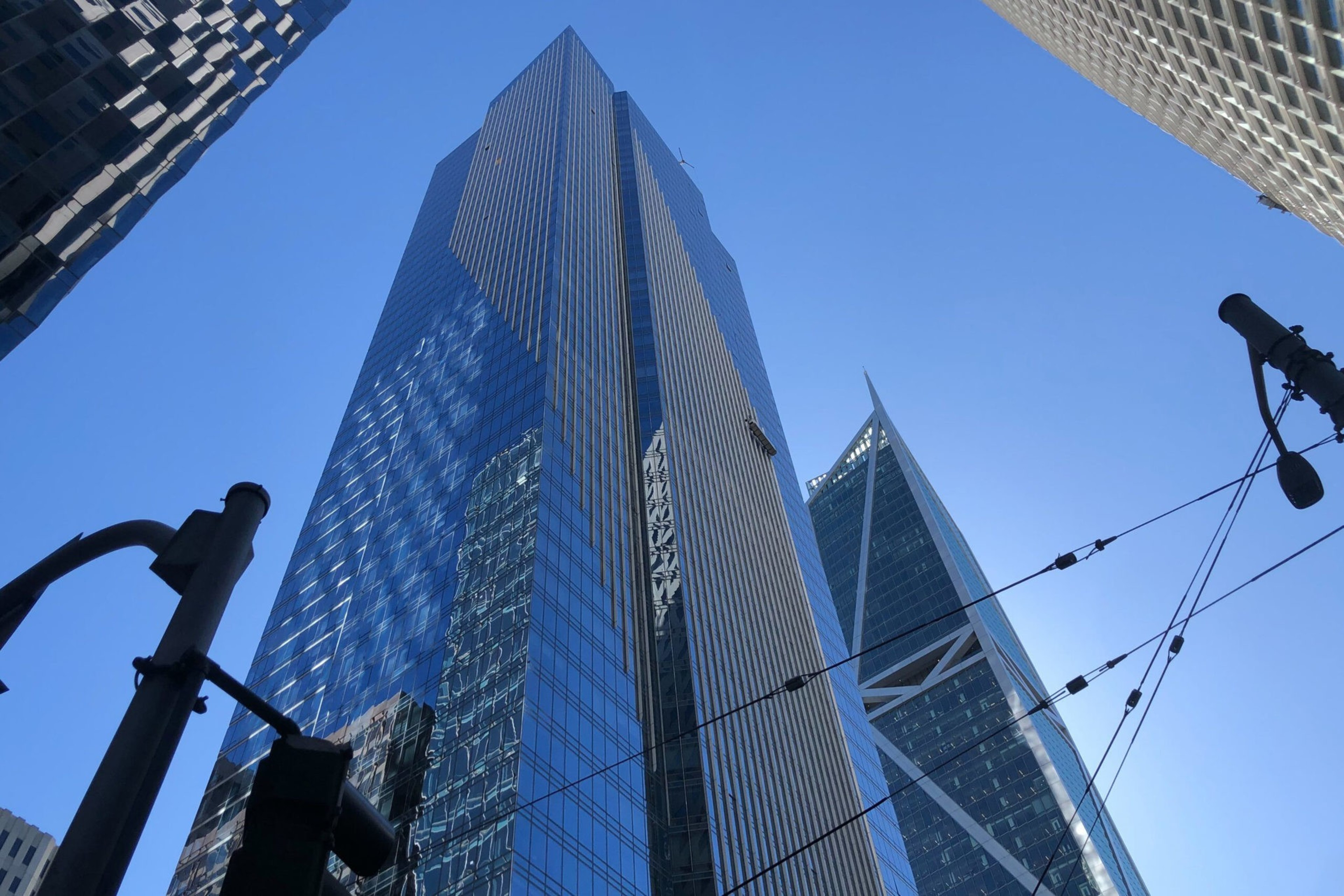 Two tall skyscrapers with glass facades reflect sunlight against a clear blue sky, with streetlight poles and overhead cables in the foreground.